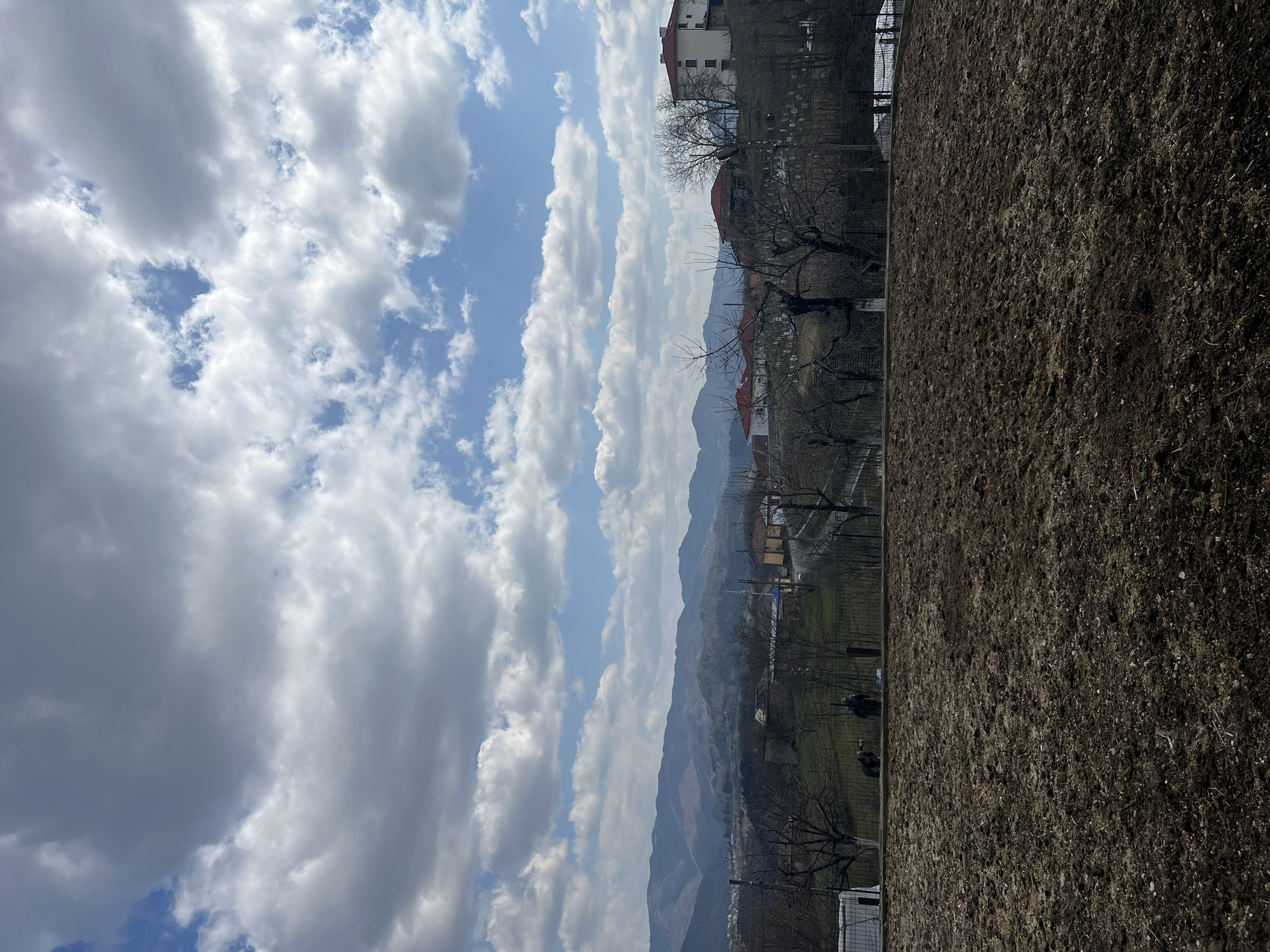 The image depicts a rural or semi-urban landscape in Sedlartsi, Bulgaria, under a partially cloudy sky. The foreground is dominated by a field of freshly tilled, dark brown soil, occupying the lower half of the frame. Directly beyond this cultivated area, a dark metal fence runs horizontally. Two individuals are visible near this fence line; one figure, dressed in dark clothing, is bent over or kneeling on the left, while another individual, also in dark clothing, stands upright slightly to their right.
Beyond the fence and a gradual slope, the village of Sedlartsi unfolds. Several multi-story residential buildings with red-tiled roofs and light-colored facades are discernible, along with sparse, leafless deciduous trees indicative of late winter or early spring. A prominent mosque with a cylindrical minaret is situated towards the center-left of the midground village view. To the right, a hillside cemetery with numerous white headstones is visible. The background features undulating mountain ranges stretching across the horizon. The sky is characterized by large, scattered white and gray cumulus clouds against patches of bright blue, suggesting daylight conditions.