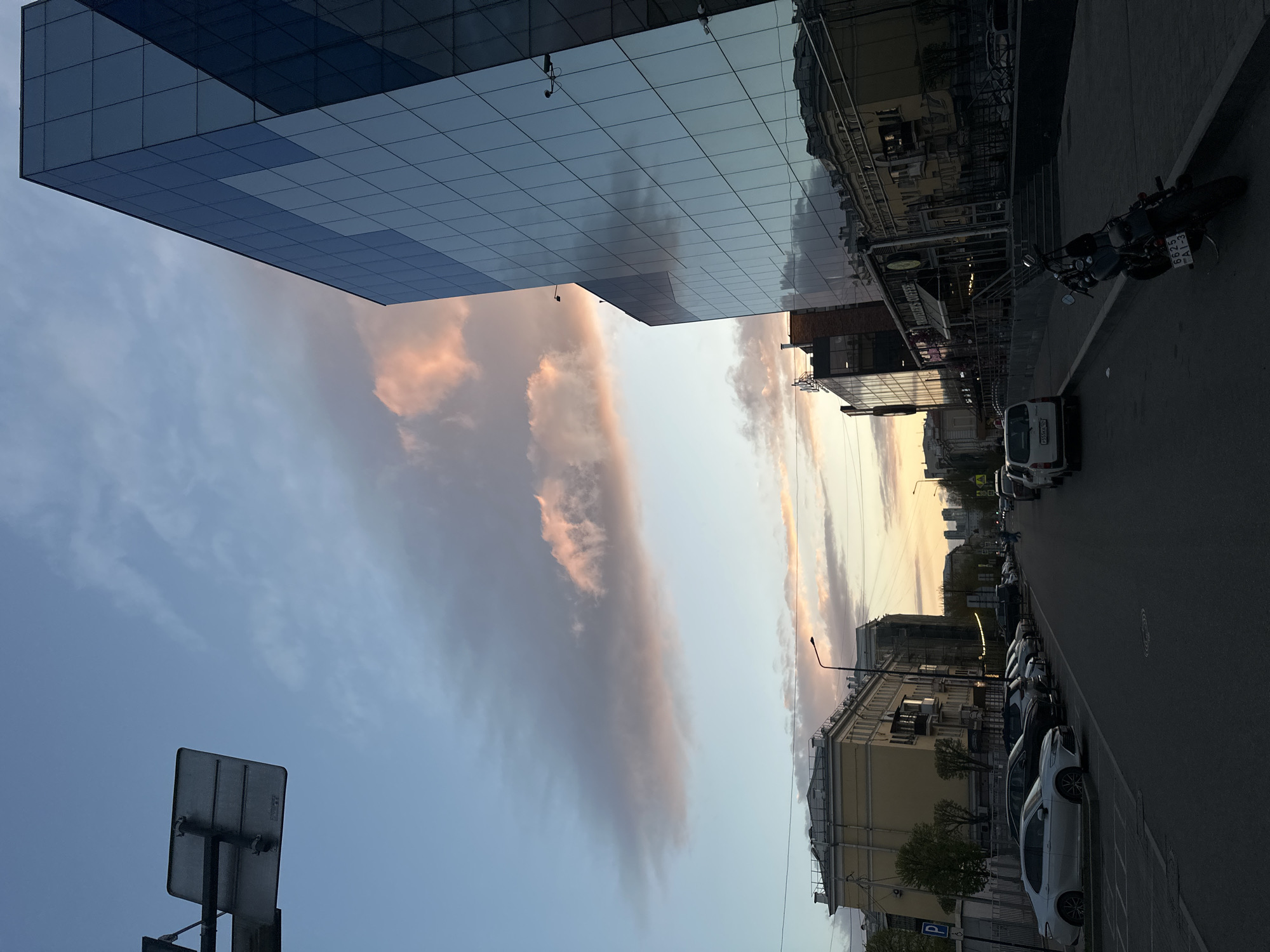 The image captures an urban street scene in Krasnoyarsk, Russia. A modern, glass-facade building dominates the left side of the frame, reflecting the sky and clouds. Adjacent to the building is a street lined with older, traditional buildings on the right. Parked cars and a motorcycle occupy the right side of the street. The sky above features elongated clouds, suggesting either dawn or dusk lighting conditions. A traffic sign is visible in the lower left corner. No people are visible in the scene.