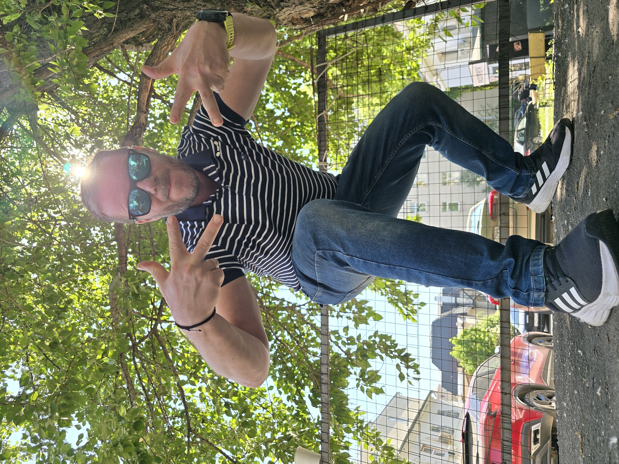 A man is standing on a street, making peace signs with both hands. He is wearing a striped shirt, blue jeans, and black and white sneakers. He has a watch on his left wrist and a bracelet on his right wrist. The man is also wearing sunglasses. Behind him is a metal fence, trees, cars, and buildings. The location may be Moscow, Russia.