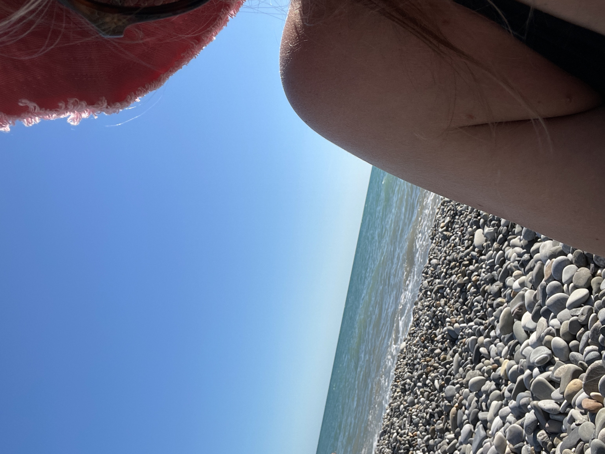 The image depicts a close-up view of a person sitting on a pebble beach. The focus is on the person's shoulder and arm, which are bare and have some fine hairs visible. To the left of the person, a clear blue sky stretches above the ocean, which has gentle waves and a white foamy edge where it meets the shore. The beach is covered in smooth, gray and brown pebbles of various sizes. In the upper right corner, the edge of a pink hat is visible, along with the person's hair and sunglasses. The scene conveys a sense of relaxation and a summer day at the beach.