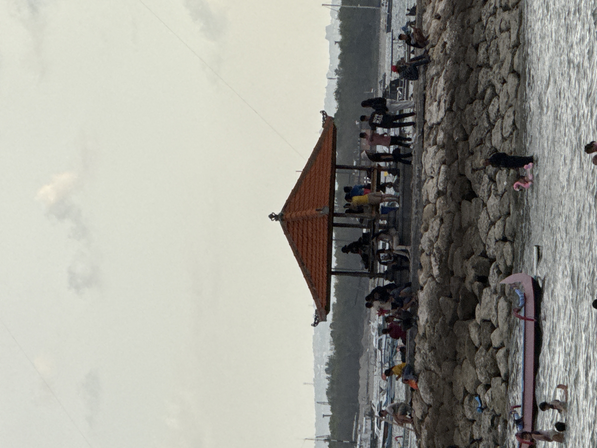 A wide shot captures a waterfront scene in Denpasar, Indonesia, likely a harbor or bay area, with a breakwater made of large rocks in the foreground. On top of the breakwater, a group of people are gathered near an open-sided pavilion with an orange tiled roof. Some are sitting on the rocks, while others stand or walk. In the water below the breakwater, there is a pink boat with people in it and a pink inflatable flamingo with a person standing near it. The background shows a treeline and several boats docked in the water, with a hazy, overcast sky above. The overall impression is a casual, public gathering place by the sea.
