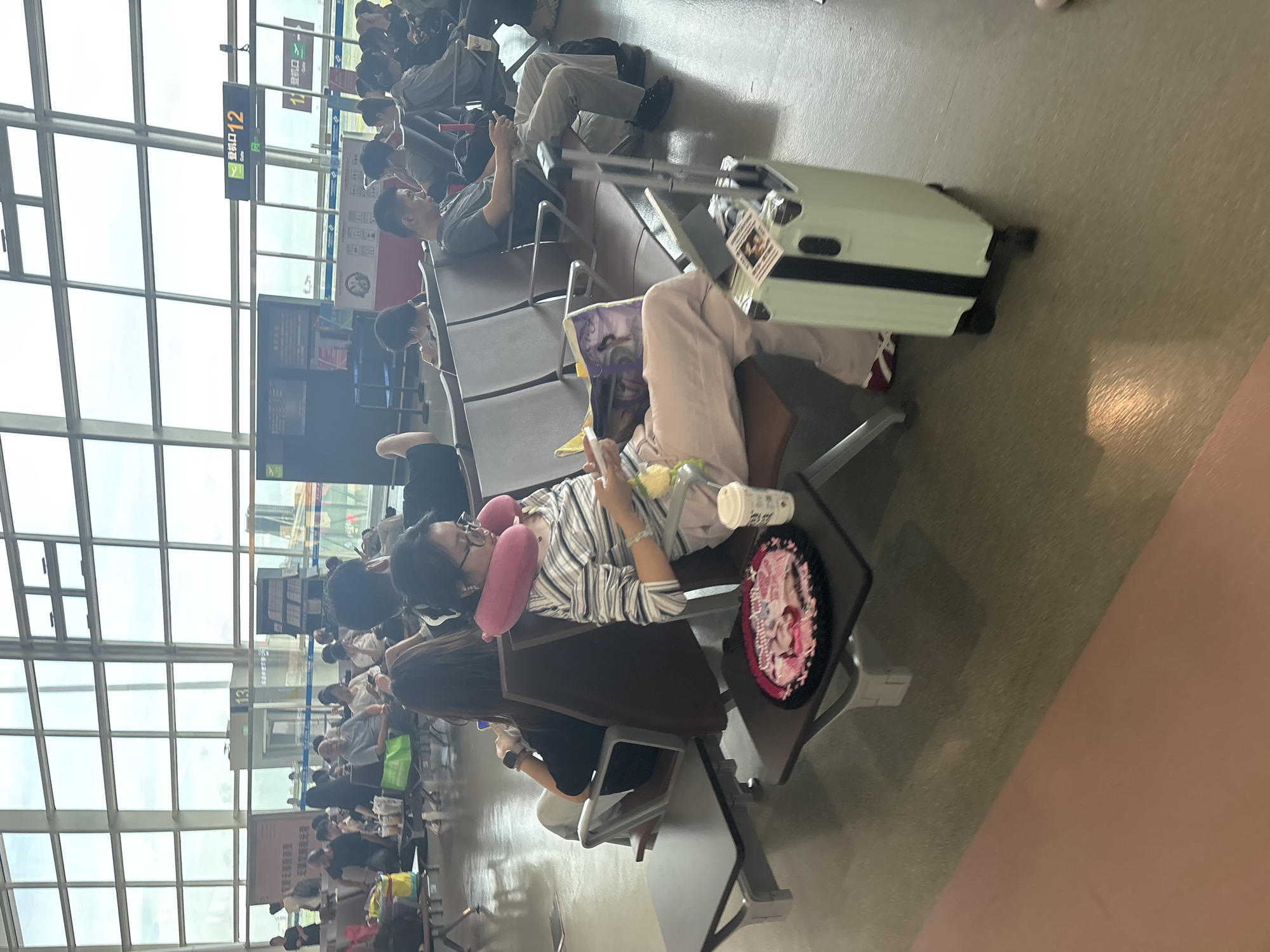 The image captures a waiting area at Xinwu District Airport in China, as indicated by the signage "登机口 12" (Gate 12). Several passengers are seated, appearing to await their flights. In the foreground, a woman with glasses and a pink neck pillow is engrossed in a book. Beside her, a light-colored suitcase with black accents stands ready. Other passengers are scattered throughout the seating area, some looking at their phones or resting. The environment suggests an airport lounge, characterized by rows of seating, large windows, and overhead signage typical of such a facility.