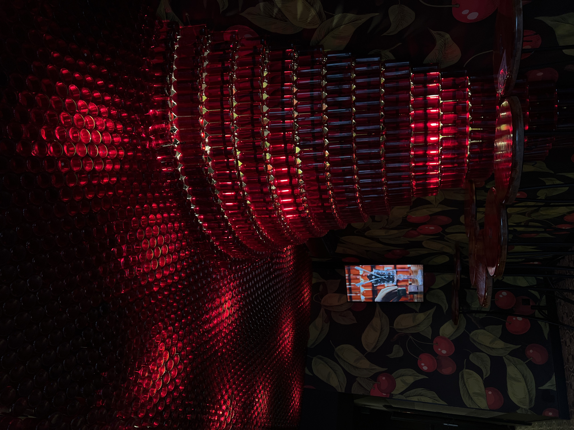 The image depicts the interior of a dimly lit establishment, likely a bar or restaurant, in Lublin, Poland. The dominant feature is a large, multi-tiered chandelier or light fixture constructed from numerous red glass bottles, casting a vibrant red glow throughout the space. The ceiling is also covered in a similar arrangement of red glass objects, creating a textured, illuminated surface. The walls are adorned with a dark wallpaper featuring a repeating pattern of cherries and leaves. Several small, round tables with red tops are visible, arranged in a semi-circular pattern. A small screen is mounted on the wall, displaying an image of a cocktail being prepared with cherries and blue straws. No people are visible in the frame.