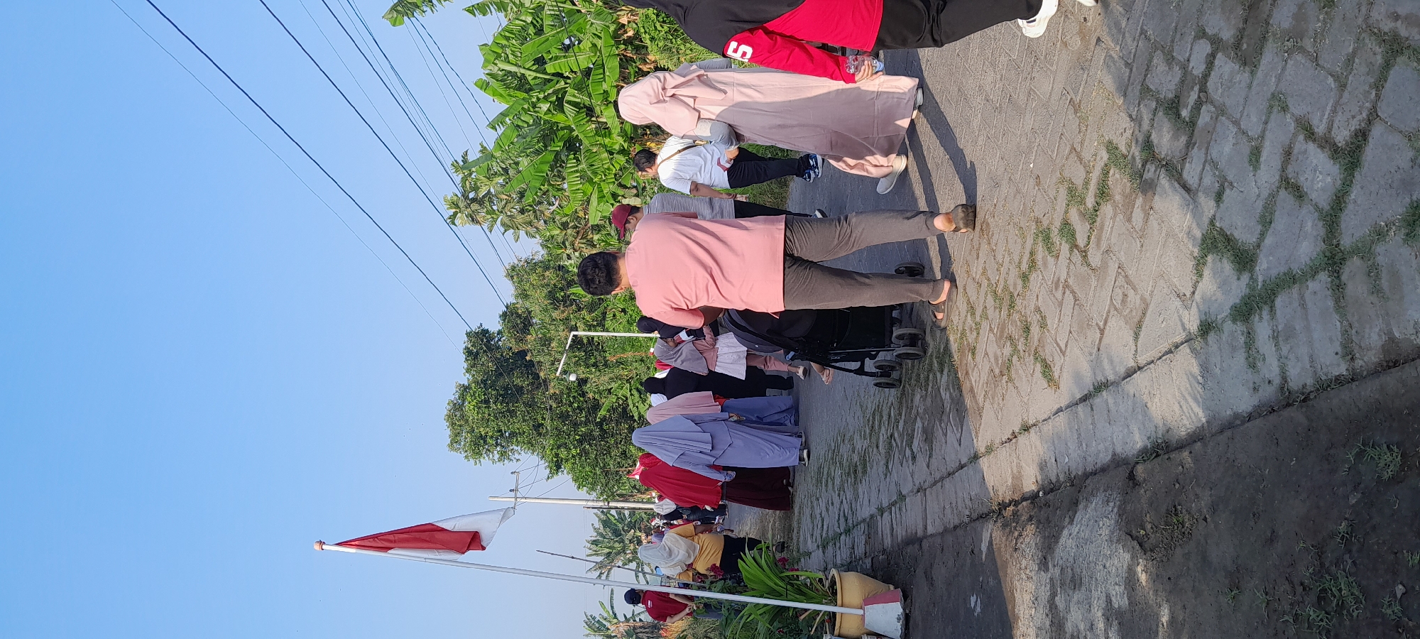 In Bendungan, Indonesia, a group of people are walking along a paved pathway lined with potted plants and trees. The sky is clear and blue, and a red and white Indonesian flag is flying on a flagpole to the left of the frame. Several individuals are wearing traditional Islamic attire, including hijabs and long dresses. One person is pushing a stroller, and another is holding a plastic water bottle. The pathway appears to be made of interlocking bricks, with grass growing between some of the gaps. The scene suggests an outdoor gathering or event taking place on a sunny day.