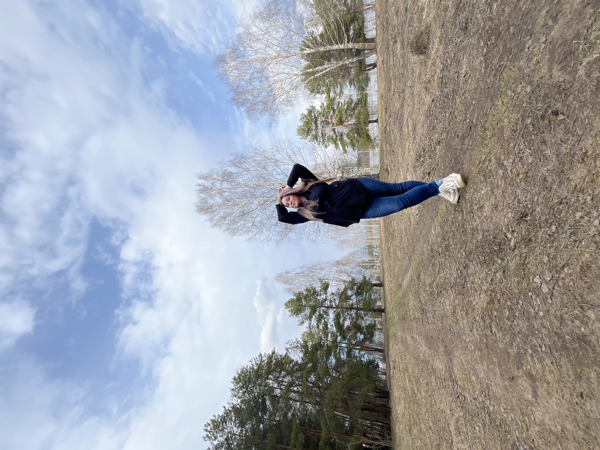 The image shows a woman lying in a field with trees and a sky with clouds in the background. The woman is wearing jeans, a black jacket, and white sneakers. She has her hands behind her head. The field is brown and dry, with some patches of grass. The trees are bare except for the pine trees. The sky is blue with white clouds. The location of the image is in the city of Chaikovsky, Russia.