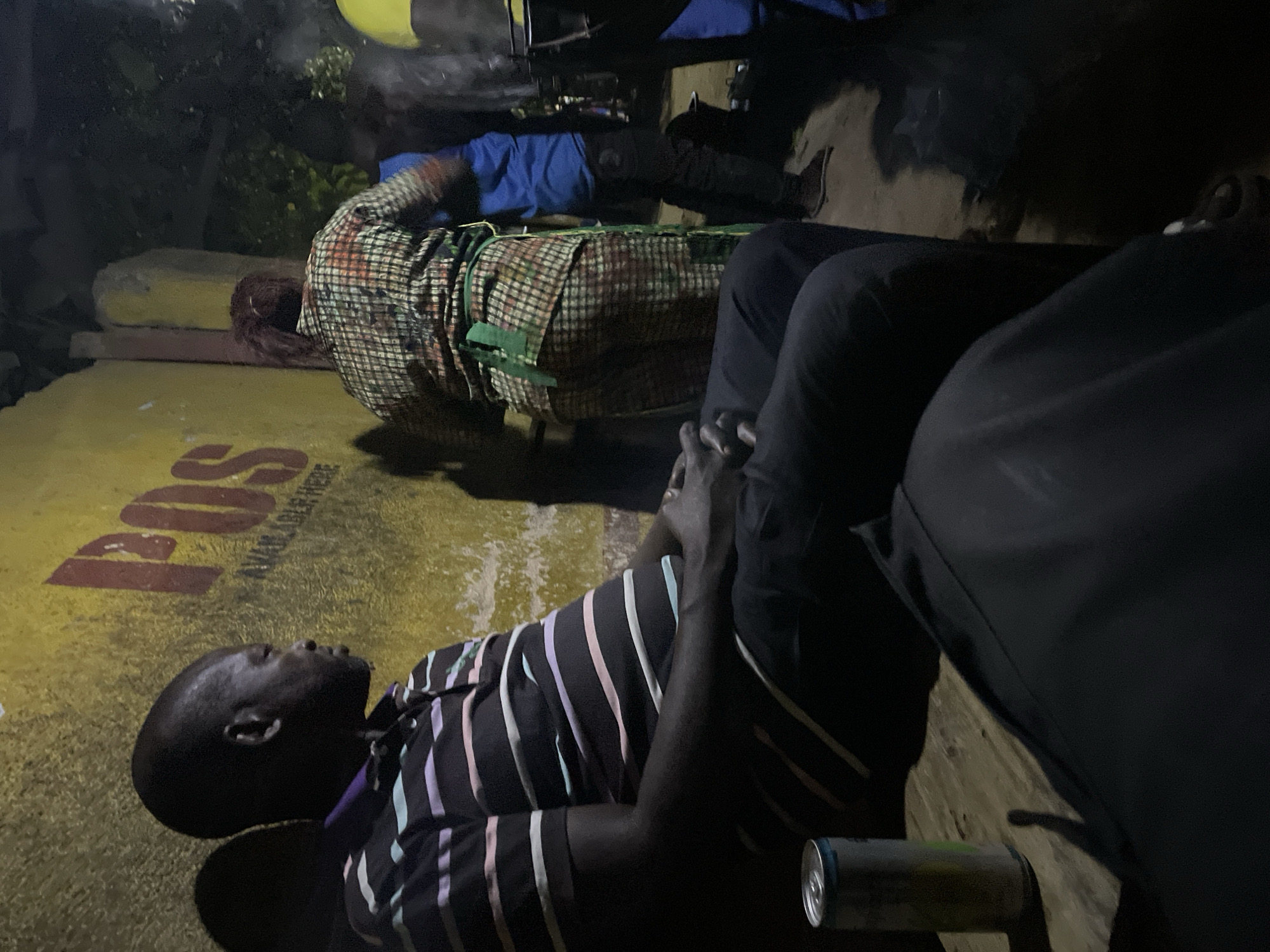 The image shows an outdoor scene at night, possibly in Jos, Nigeria, given the presence of the word "JOS" on a yellow surface. A man wearing a striped shirt is lying on the ground. There is an unopened drink can laying near him. To his right, a person wearing black pants is sitting. Behind him is another person lying on a yellow surface, covered with a checkered blanket and blue fabric.