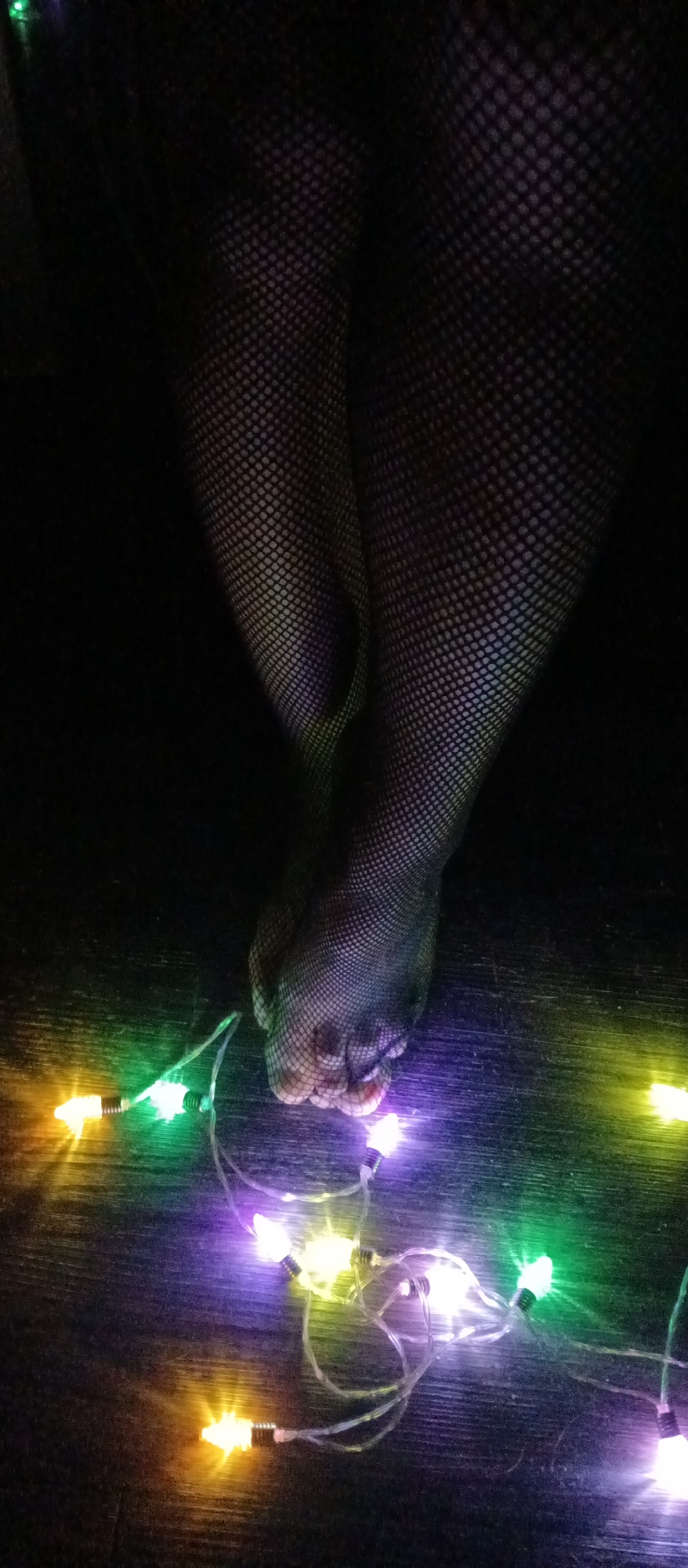 A close-up shot shows two feet clad in black fishnet stockings on a dark wooden surface. Christmas lights, radiating colors of green, yellow, and purple, are intertwined around the toes. The photograph is taken in a location of unknown, unknown.