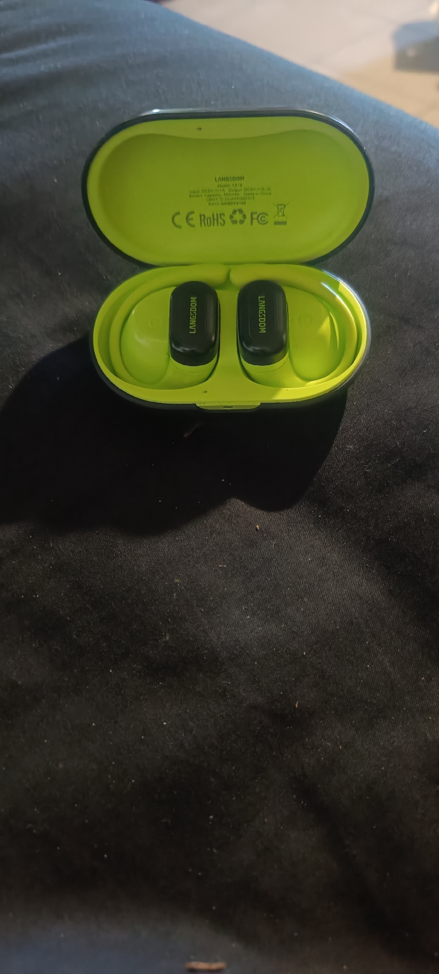 This image shows a pair of LINGSCOM wireless earbuds in a bright green charging case, which is open. The earbuds themselves are black and have "LINGSCOM" printed on them in white text. The inside of the charging case also has text printed on it, including "LINGSCOM", "Model: 1819", "Input: DC5V 1A Output: DC5V 1A", "Battery Capacity:", "Made in China", "FCC ID: 2AJ4XOB2312", and "Batch ID: BMBFP0103". The case also displays CE, RoHS, and FC certification marks, as well as a symbol indicating it should not be disposed of with household waste. The charging case is placed on a dark, textured surface that appears to be fabric. The image is shot from a slightly overhead angle, with a shadow cast by the earbuds and case onto the surface below. There are no people or animals visible in the image, and the environment suggests an indoor setting. The location is identified as Semenivka, Ukraine.