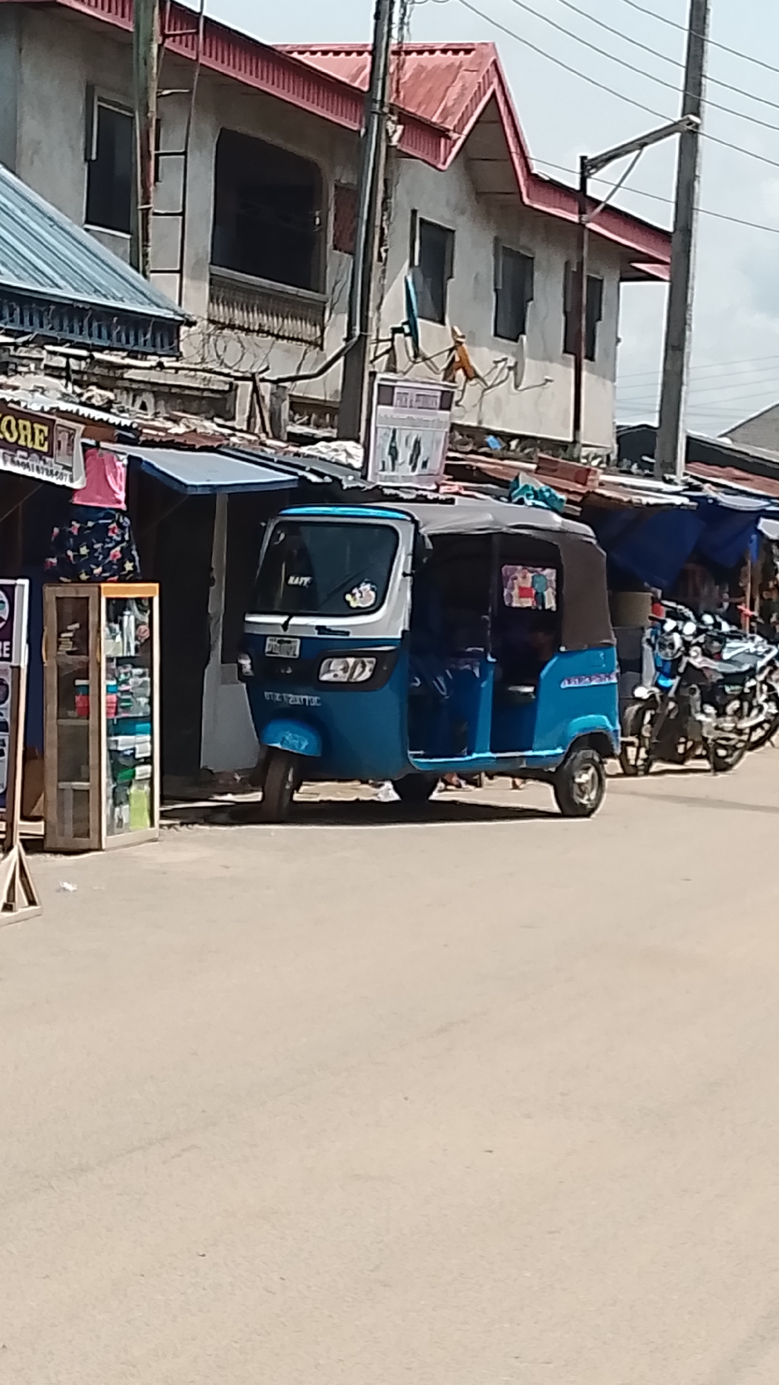 A blue and white auto rickshaw is parked on a street in an urban area, likely a developing country. The vehicle has a black canvas top and is positioned in front of a row of small shops with blue awnings. Several motorcycles are parked nearby. The buildings in the background appear to be residential and commercial structures with a mix of corrugated metal and traditional roofs. Power lines and utility poles are visible above the structures. The street surface is a light-colored material, potentially concrete or asphalt.