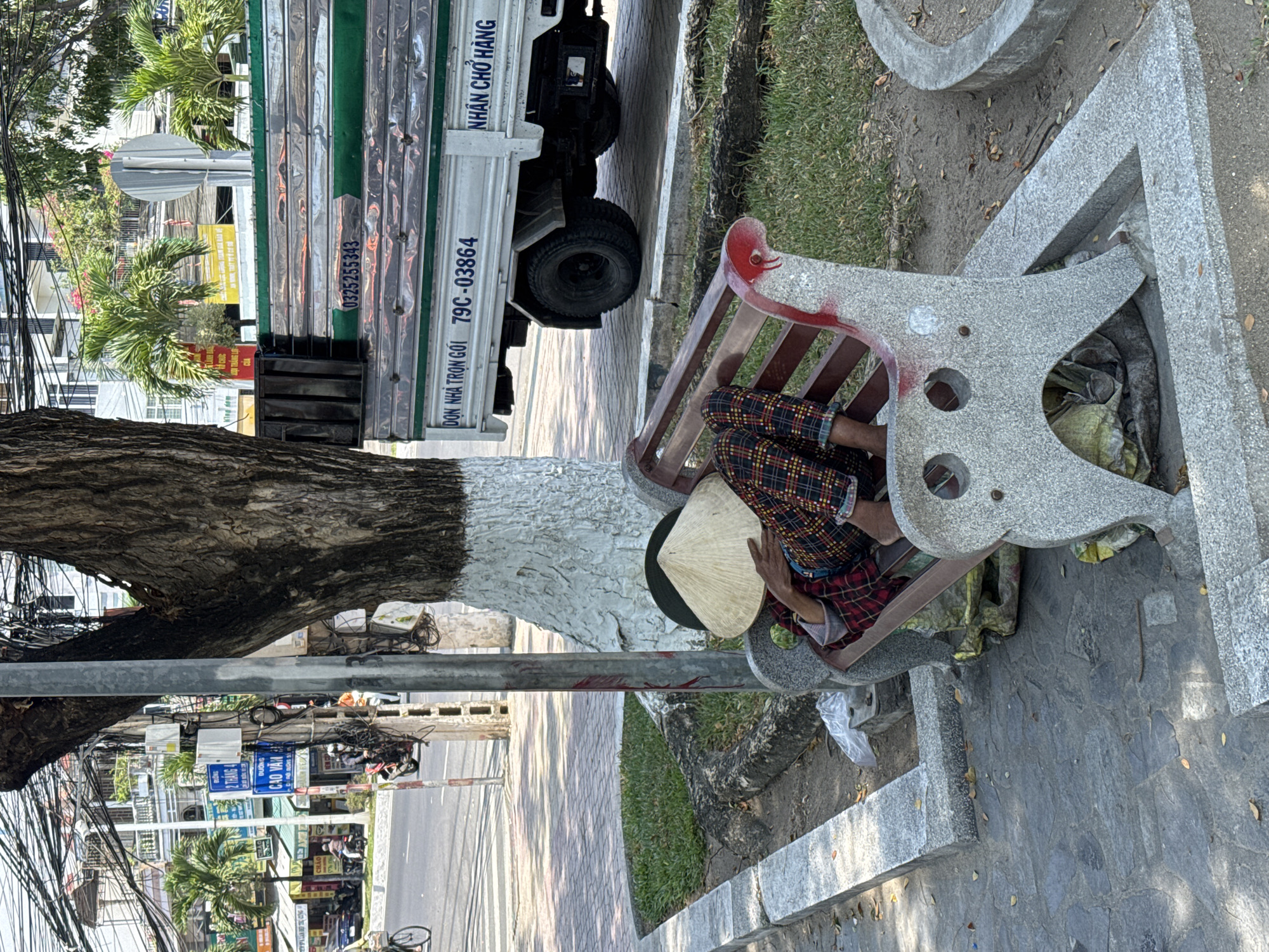 A person wearing a conical hat and plaid pants is sleeping on a park bench in the city of Bac Nha Trang Ward, Vietnam. A large tree with a painted white trunk is next to the bench. A truck is parked on the street adjacent to the park. Street signs for "Duong Cao Van" and "2 Thang 4" are visible in the background. The truck has the phone numbers "0325255343" and "79C-03864" printed on its side. The phrase "DON NHA TRON GOI NHAN CHO HANG" is also printed on the truck. The park area has grass, trees, and paved walkways. Overhead power lines are visible.