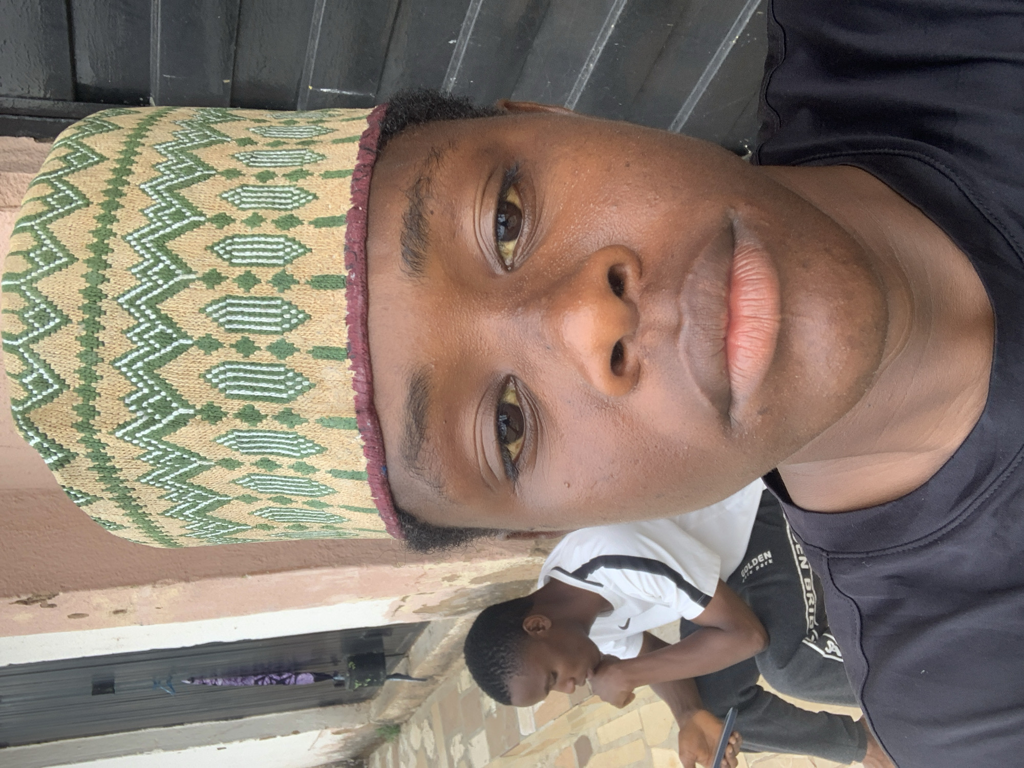 A medium-close-up, sideways-oriented photo shows a Black man with dark eyes, wearing a decorated tan, green, and white hat with a maroon edge. He has dark hair and is wearing a black T-shirt. Behind him and to the side, a younger Black boy is wearing a white T-shirt with black stripes on the sleeves and dark pants. The younger boy is seated and looking down. In the background, there is a brown wall and a black, textured surface. Based on visual cues, the location is possibly Jos, Nigeria.