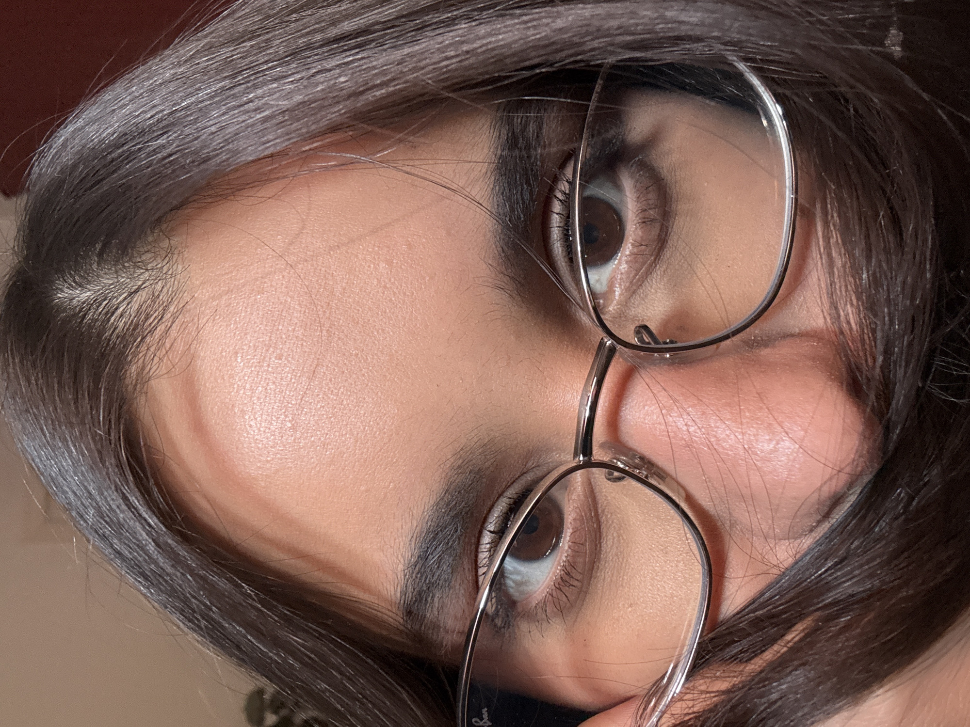 The image is a close-up of a person's face, focusing on their eyes and forehead. The person is wearing eyeglasses with thin metal frames. Their dark hair is partially covering their face and frames. The skin tone appears to be light to medium. The background is blurred and out of focus, with warm, muted tones. There are no discernible elements in the image that would suggest a location in Las Rozas de Madrid, Spain.