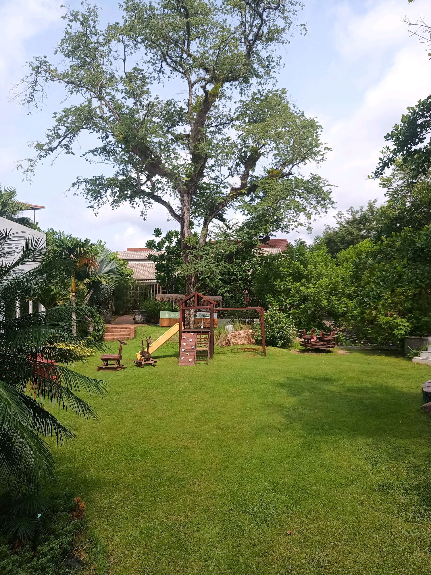 The image depicts a lush green backyard playground area with a large, mature tree dominating the background. In the foreground, a well-maintained lawn is visible, with a wooden playground set featuring a yellow slide and a climbing wall. Two small wooden animal-shaped benches are placed on the grass in front of the playground. Further back, a covered structure with a thatched roof, possibly a seating area or pavilion, can be seen. The area is surrounded by dense foliage and trees, with a building with a tiled roof visible behind the playground. The sky is partly cloudy. The location appears to be Tambon Pak Nam, Thailand, given the tropical vegetation and architectural style. There are no people visible in the image.