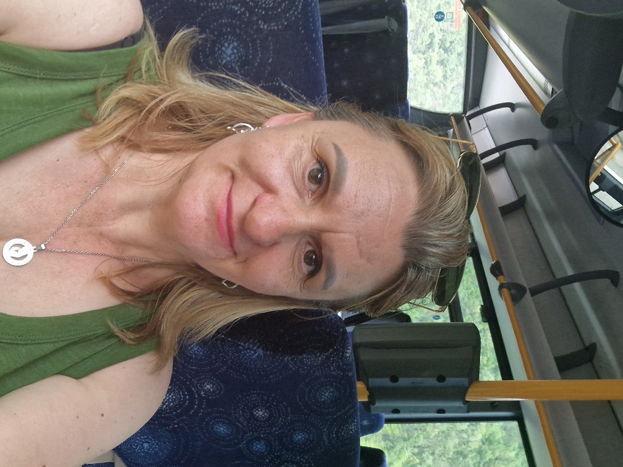 This is a selfie taken from a seated position inside a bus. The subject is a woman with blonde hair and light skin, wearing a green tank top and a silver necklace with a pendant. She is looking directly at the camera with a slight smile. Sunglasses are perched on her head. The background shows the interior of the bus, including blue seats with a patterned design, and windows through which trees and foliage are visible. The overhead interior of the bus is also visible, with yellow poles and handles. The location is not specified as Verrès, Italy.