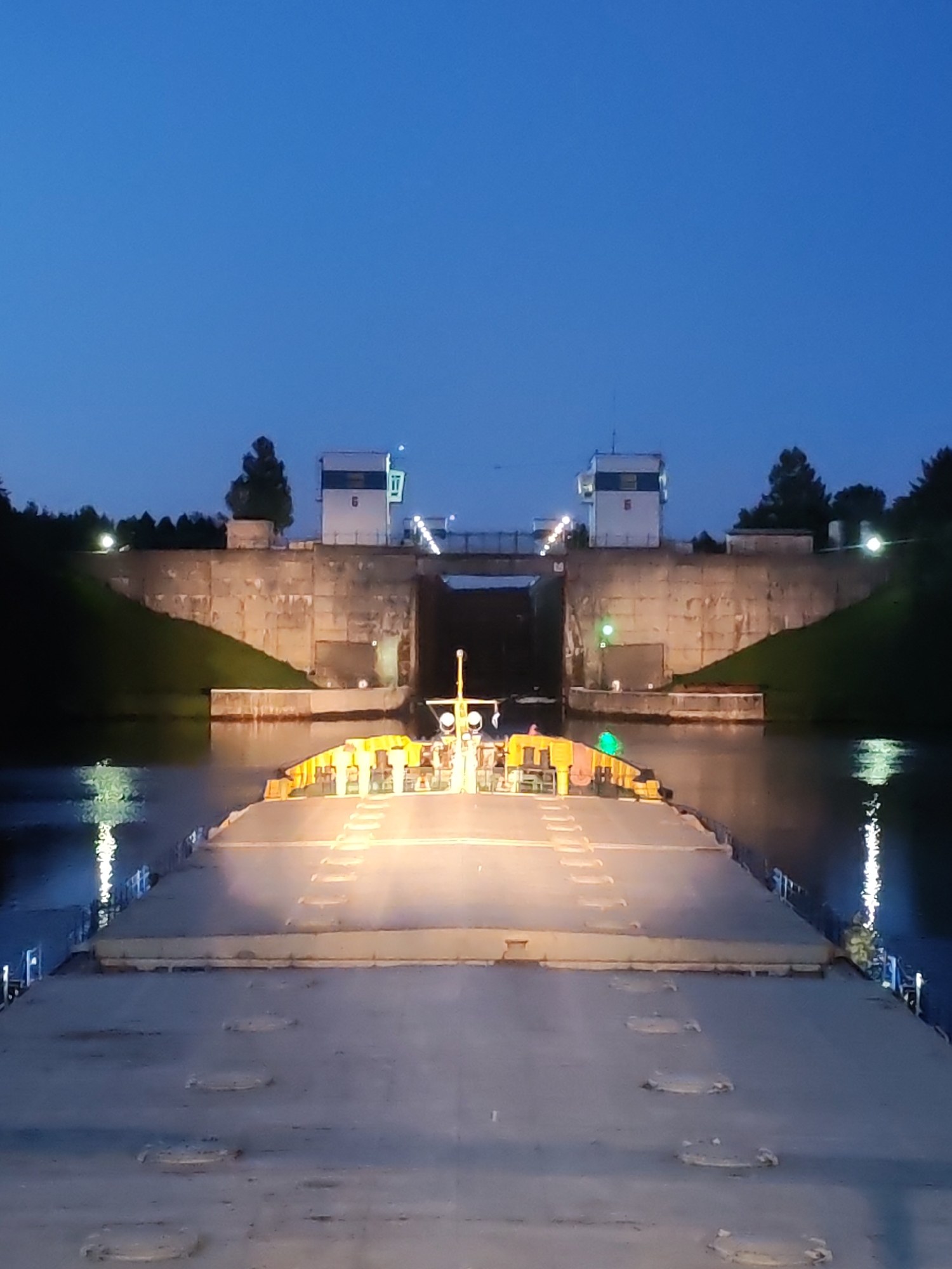 The image captures a nighttime or late dusk scene of a large vessel, likely a barge or ship, approaching a canal lock. The perspective is from the deck of the vessel, looking forward towards the lock entrance.
The vessel's flat deck, visible in the foreground, shows several circular hatches and a marked walkway. The forward part of the vessel is brightly illuminated by yellow/orange lights, possibly from crew quarters or equipment, with a mast-like structure also adorned with lights.
In the distance, the concrete structure of the lock is prominent. It features two white control towers, one on each side, perched atop the concrete walls. The right tower clearly displays the number "6" on its side, possibly indicating the lock number. The lock entrance is illuminated by bright lights, and a green light signal is visible, perhaps indicating clearance to proceed. Grassy banks flank the concrete walls of the lock, and dark trees are silhouetted against the sky on both sides.
The sky is a deep, clear blue, characteristic of twilight or nighttime, with no visible clouds. The water in the canal reflects the artificial lights from the vessel and the lock, creating shimmering patterns. The overall impression is one of calm, nocturnal navigation through an engineered waterway. The location context provided is Devyatiny, Russia, a region known for its extensive canal systems.