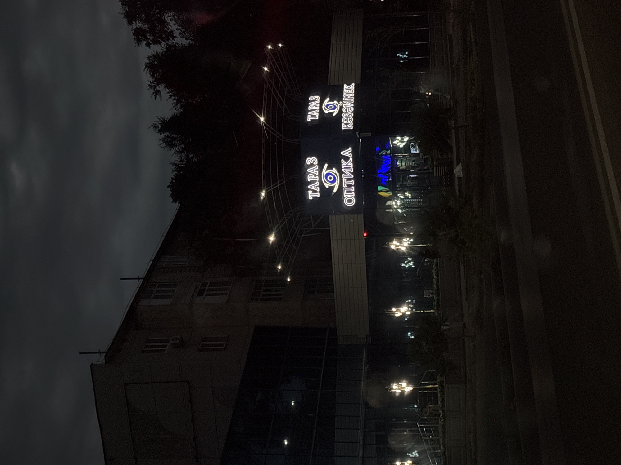 The image captures a nighttime view of a building with prominent neon signs, likely in Taraz city, Kazakhstan. The signs display text in what appears to be Cyrillic and possibly another script. One sign reads "ТАРАЗ ОПТИКА" with an eye symbol, and another reads "ТАРАЗ КӨЗЭЙНЕК" also with an eye symbol. The building itself seems to be a multi-story structure with a dark facade and illuminated windows. The scene is lit by the neon signs and streetlights, creating a somewhat atmospheric and urban nighttime setting. The overall impression is one of a commercial establishment, possibly an optical shop, at night. The weather appears overcast, contributing to the dark mood of the image. There are no people clearly visible in the frame.