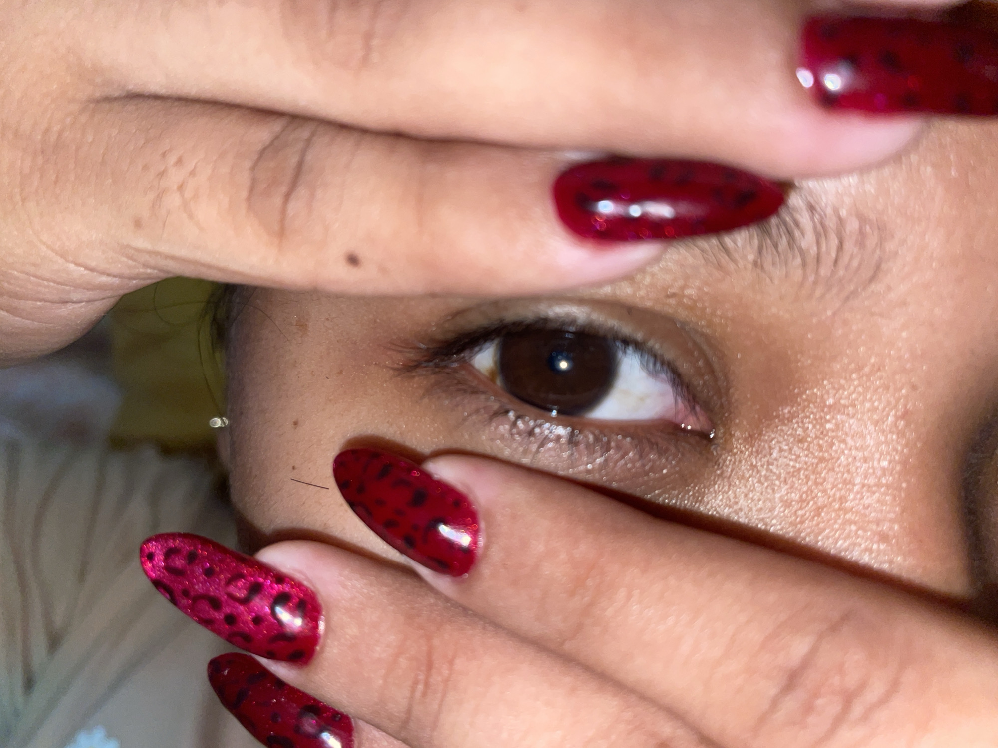 A close-up shot depicts a person's eye partially obscured by their hands. The fingernails are painted a deep red with black leopard print designs. Skin tone appears to be medium brown. A single tear glistens in the corner of the eye. A small mole is visible near the eyebrow. The background is blurred. The city and state are unknown.