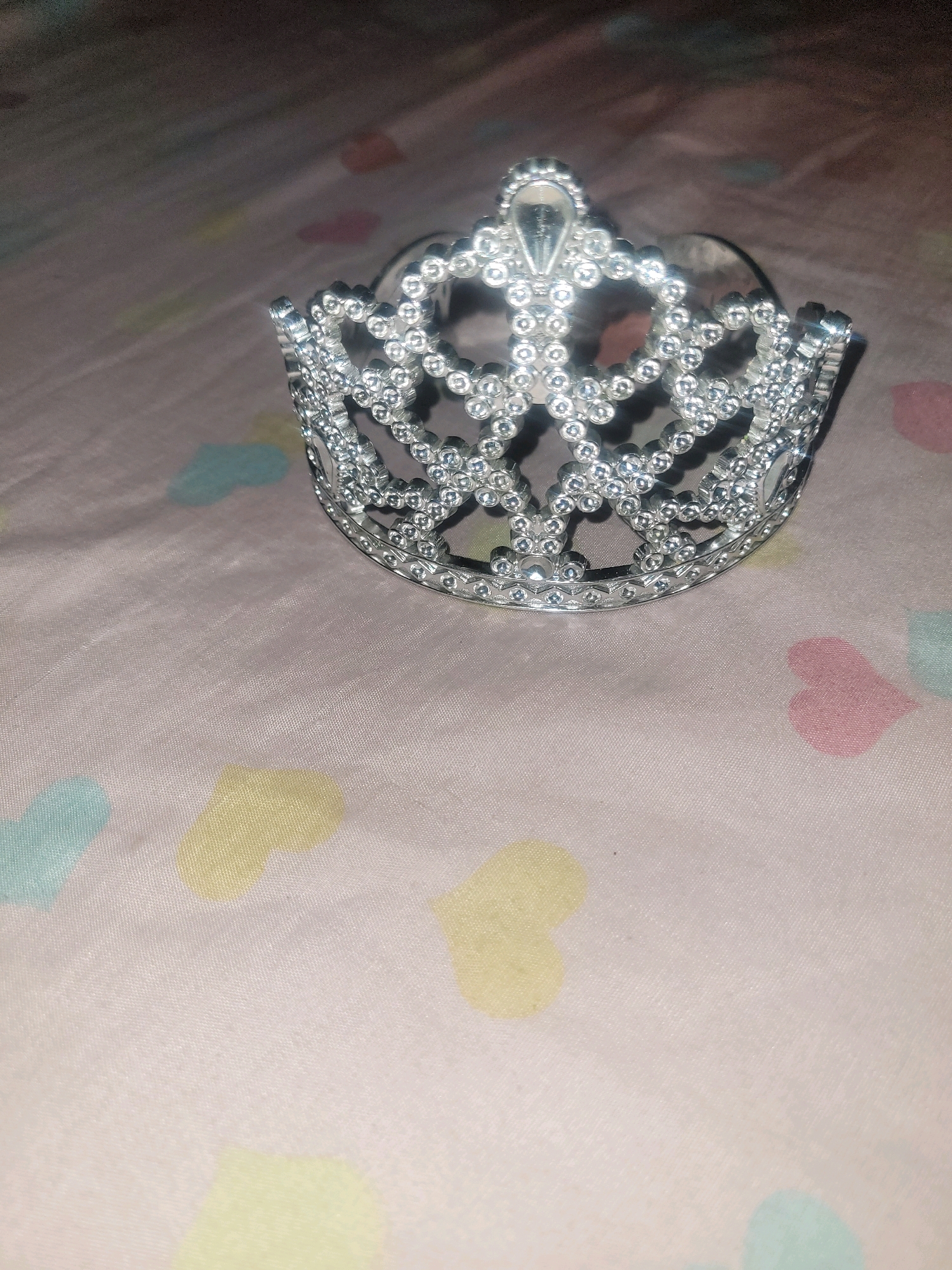 A silver tiara with a teardrop-shaped gem at its center rests on a light-colored fabric. The fabric has a pattern of pastel-colored hearts scattered across its surface, with individual hearts in shades of pink, yellow, and light blue visible. The tiara is positioned in the upper right quadrant of the image, angled slightly towards the viewer. The lighting in the image is dim, casting soft shadows and creating a slightly blurred effect, particularly on the fabric. The background is out of focus, obscuring any specific details of the environment. It is not possible to determine the location in Beausejour, Saint Lucia, from the provided image.