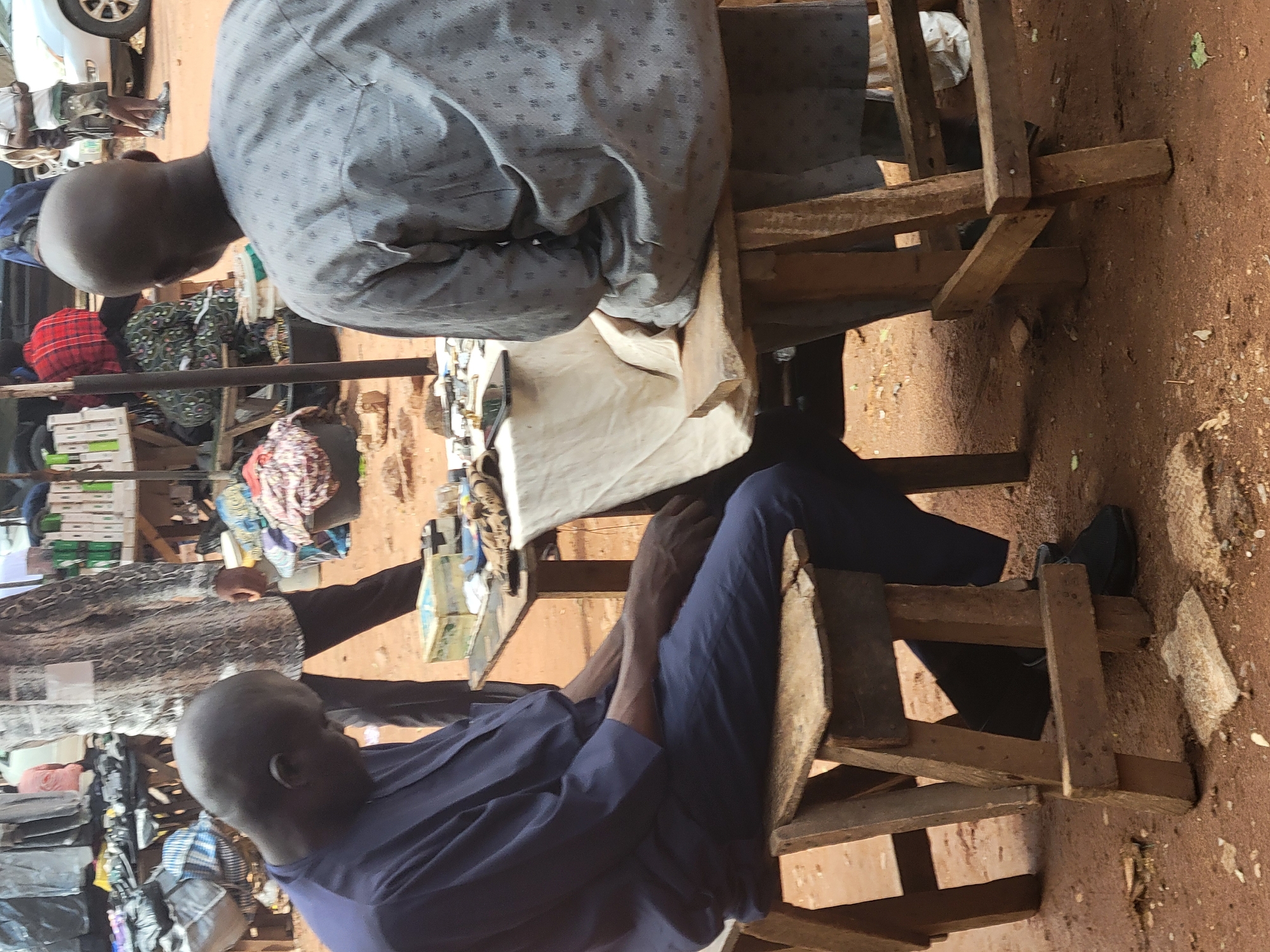 Two men sit on wooden chairs on a red-brown dirt surface in an outdoor market. One man wears a patterned grey shirt and the other a blue long-sleeved shirt and black shoes. They sit at a table with goods displayed on it. Other people and merchandise are visible in the background. The location is Abuja, Nigeria.