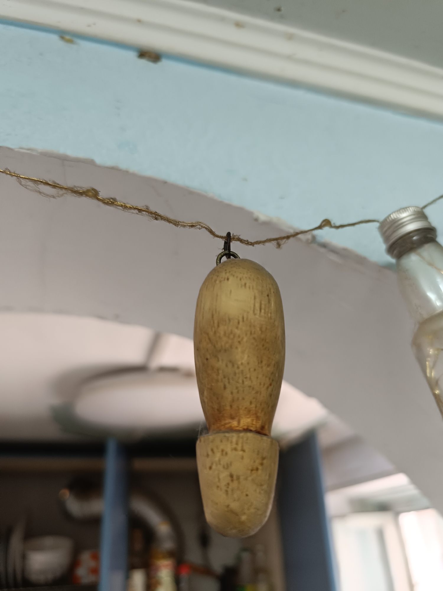 A close-up, low-angle shot shows a wooden object hanging from a thin, gold-colored wire. The wooden object is roughly cylindrical with a rounded top and a narrower, flared bottom. It appears to be a decorative element or a part of a larger item. The wire is strung across an archway. To the right of the wooden object, a glass bottle with a silver cap is also hanging from the wire, appearing to be part of the same decorative string of objects. The background is out of focus, showing a light blue wall and a white ceiling. Below the archway, a shelf unit with various items, possibly in a kitchen or storage area, is visible. There is no indication of the location being in Taraz city administration, Kazakhstan.
