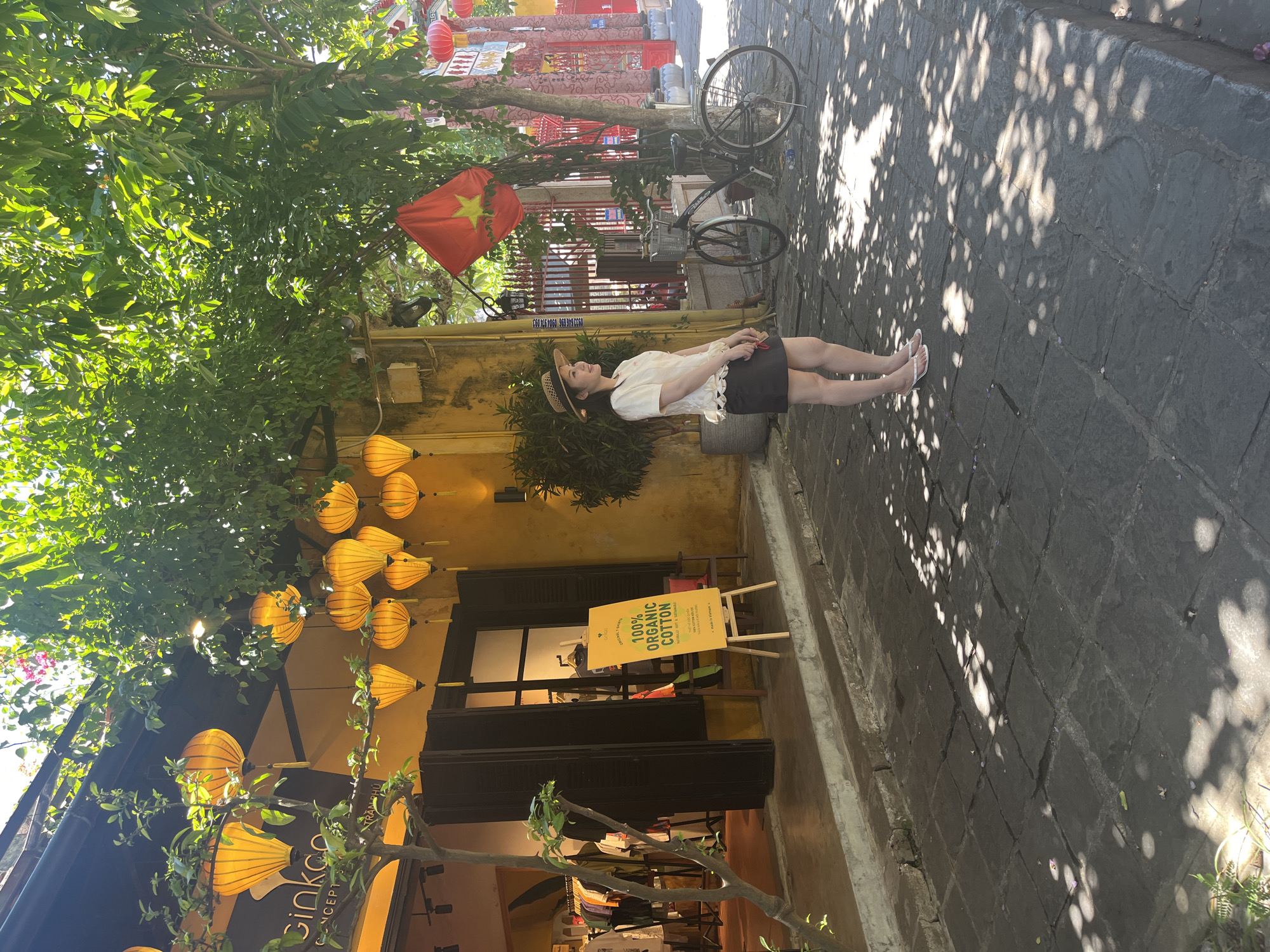 This media file captures a vibrant outdoor street scene, likely in Hoi An, Vietnam, given the traditional architecture and prominent lanterns. The setting is a paved stone street flanked by old buildings with yellow-painted facades and lush green trees providing shade.
The central subject is a woman standing on the right side of the street. She is wearing a white short-sleeved top with ruffles, a dark skirt, a straw hat, and white flip-flops. Her head is tilted slightly upwards and to her left, seemingly observing the surroundings or the sky. Her posture appears relaxed.
Notable objects include numerous yellow traditional lanterns hanging from the building on the left, adding a characteristic cultural element to the scene. A red Vietnamese flag with a yellow star is visible hanging amidst the tree foliage above the woman. In the background, a dark bicycle is parked against a wall. The street surface is made of dark, textured stone blocks, and dappled sunlight filtering through the trees creates distinct shadows on the ground.
The bright sunlight and clear shadows indicate a sunny daytime, likely morning or late afternoon.
Visible text includes a yellow A-frame sign near a storefront that clearly reads "100% ORGANIC COTTON." Below this, smaller text mentions "ECO-FRIENDLY," "SUSTAINABLE," "HANDMADE," and "MADE IN VIETNAM." The storefront itself also shows "GINKGO CONCEPT." Faint text on a distant wall includes a phone number, "090 999 5556." The bicycle has "TREK" faintly visible on its frame.