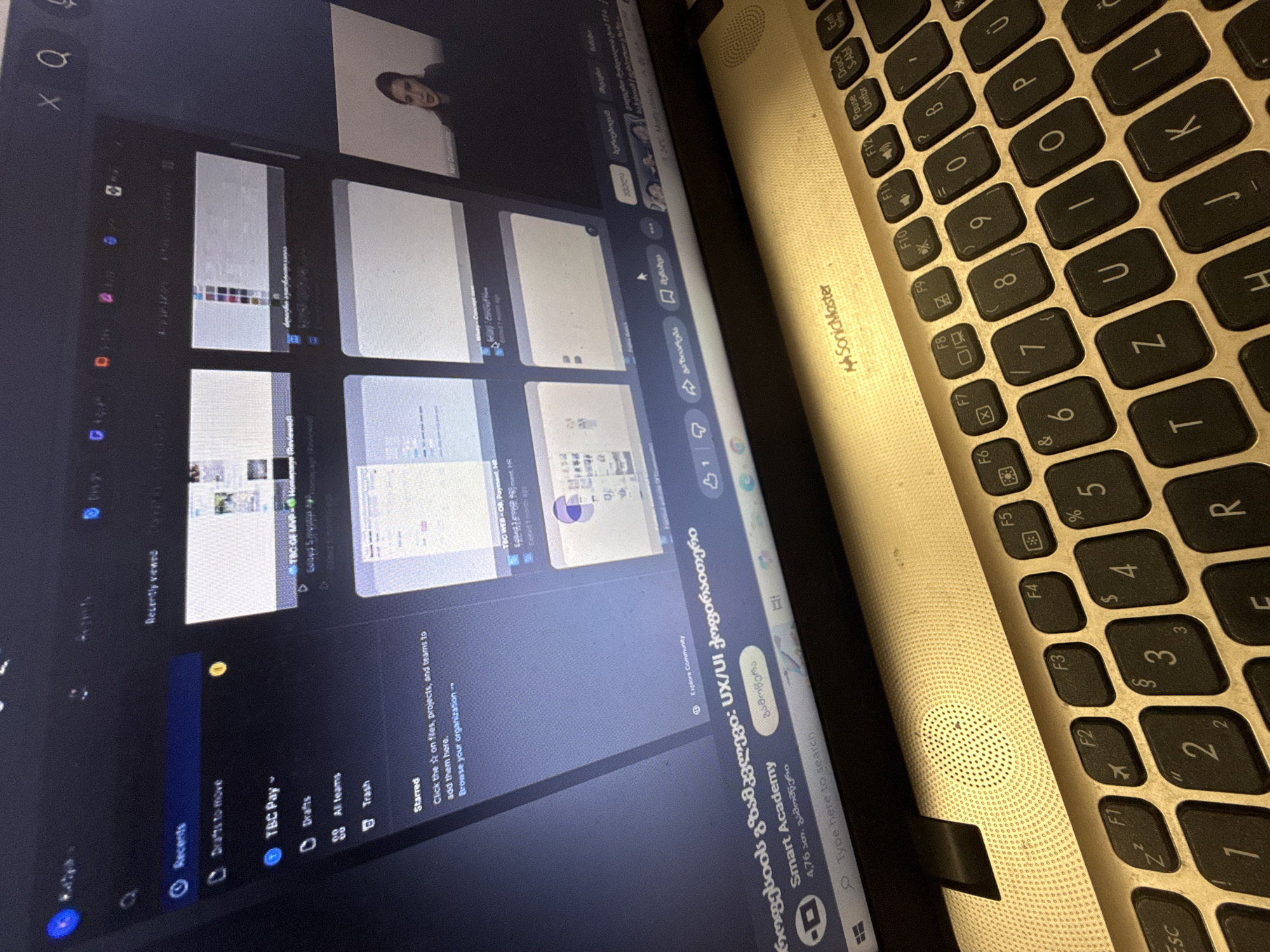 The image shows a close-up of a laptop screen and keyboard. The laptop is open, and the screen displays a website with various design projects and a video call interface. In the video call, a woman with dark hair pulled back is visible, speaking. The website interface includes tabs like "Recents," "Drafts to move," "TBC Pay," "Drafts," "All teams," and "Trash." Several project thumbnails are displayed, with titles and brief descriptions. On the lower part of the screen, Georgian text is visible, along with a search bar. The keyboard is a standard QWERTY layout, and some keys are illuminated. The overall environment appears to be indoors, with the lighting reflecting off the screen and keyboard.