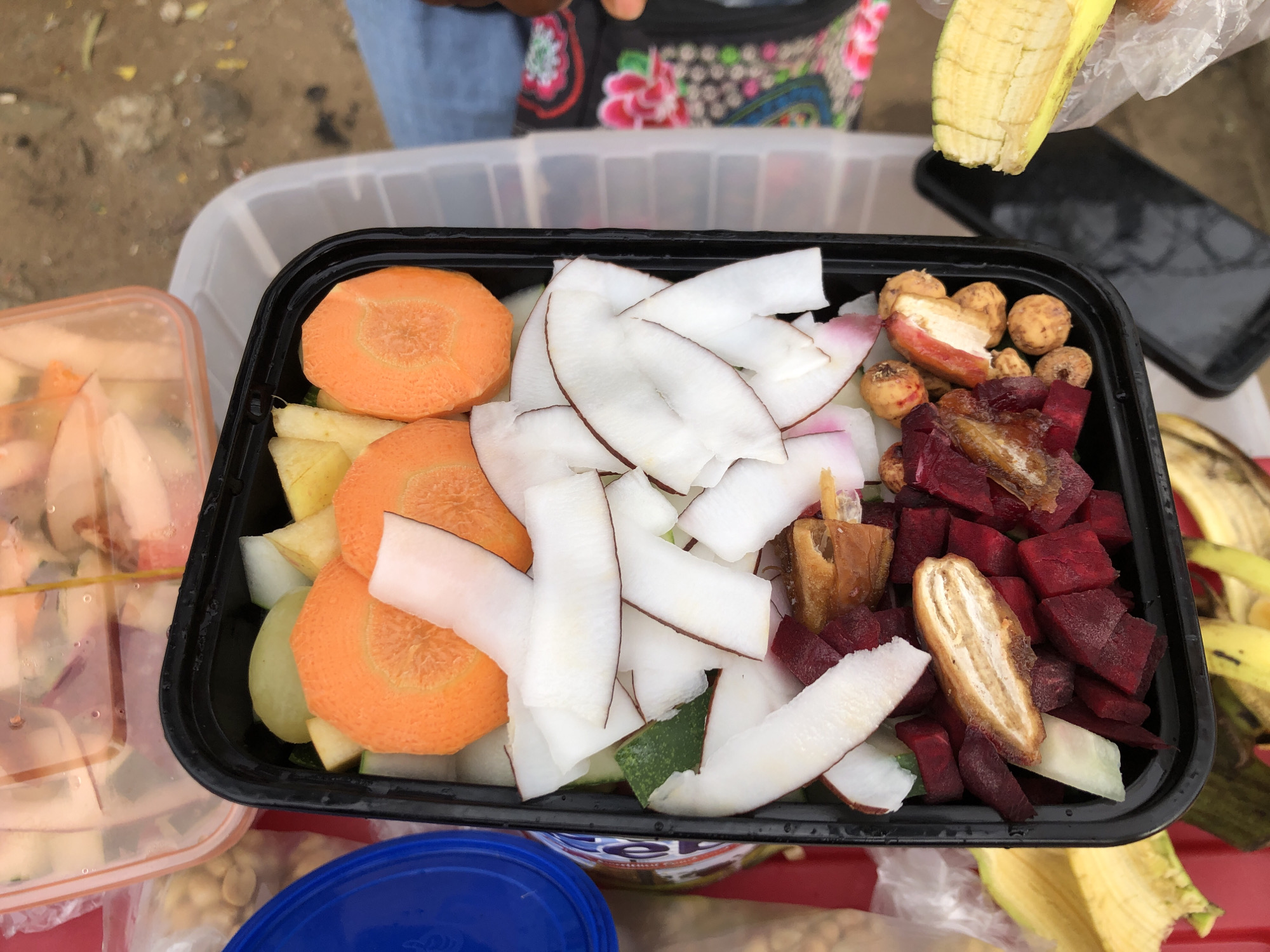 An overhead shot shows a black plastic food container filled with various fruits and vegetables. The container holds sliced carrots, white coconut strips, chopped beets, and what appear to be dates. Some small round objects, possibly tigernuts, and pieces of fruit or vegetable are also present. The container is placed on top of a larger, clear plastic container. To the left of the black container is a blue lid, and next to it, a clear plastic bag filled with peanuts. In the background, another clear plastic container holds sliced fruit. To the right, a person's leg wearing blue jeans and a colorful patterned fabric are visible. A partially peeled banana is being held by an unseen hand. The scene appears to be outdoors, with the ground covered in gravel and dirt. The overall impression is of a street-side food stall or market in Ikeja, Nigeria.
