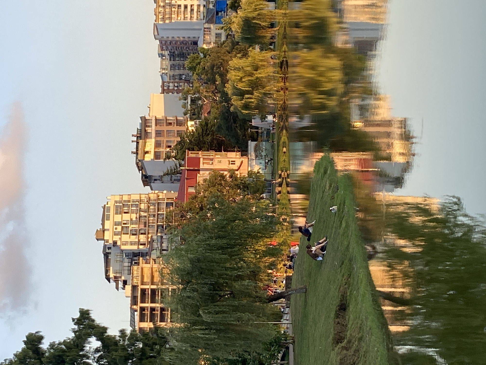 This is an eye-level, medium shot of a lakeside park in Batumi, Georgia, during sunset. The sun casts a warm glow on the buildings across the lake, which are a mix of modern high-rises and older, more decorative structures. The buildings are reflected in the still water of the lake, creating a mirror image. In the foreground, a grassy slope leads down to the lake. Several people are scattered across the slope, enjoying the evening. Some are sitting, some are lying down, and others appear to be walking. A weeping willow tree dominates the left side of the frame, its branches reaching towards the ground. The overall atmosphere is serene and peaceful.