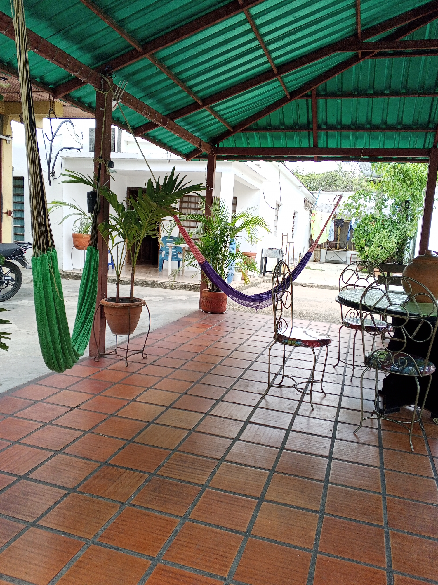 The image shows a covered patio area in Tinaquillo, Venezuela. The patio has a red-tiled floor and a green corrugated metal roof supported by brown beams. A green hammock hangs from the left side of the patio, near a motorcycle parked outside. Two potted palm trees are placed near the hammock. A purple hammock is strung between two beams further back on the patio. A wrought iron table and three chairs are arranged on the right side of the patio. Through the open end of the patio, a white building is visible, along with a clothesline in the distance.
