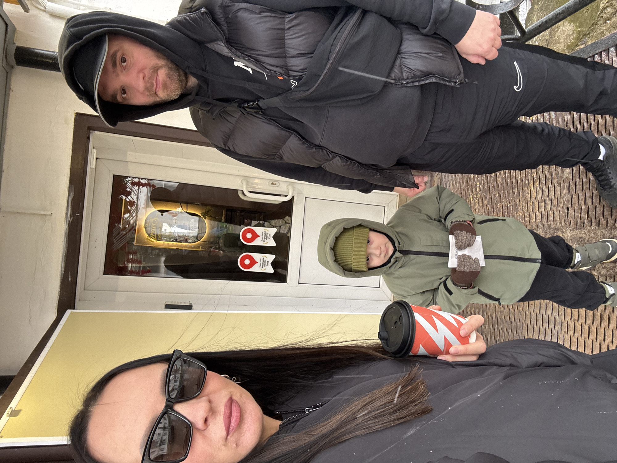Three people stand on a textured metal surface outside a building with a light yellow facade. The woman in the foreground is holding a red and white paper cup with a black lid. She wears black sunglasses and a black jacket. Behind her and slightly to the left, a child in a green hooded jacket and a green beanie holds a small paper containing what appears to be two chocolate pastries. An adult male in a black hooded jacket and black pants is standing to the left of the child, leaning slightly towards the camera. The building behind them has a white-framed glass door or window with two red circular stickers bearing white text. The window reflects the interior of the building, which appears to be a cafe or restaurant. The location is the city of Chaikovsky, Russia, as indicated by the text on the stickers "Хорошее место" and "Лучшее место 2025".