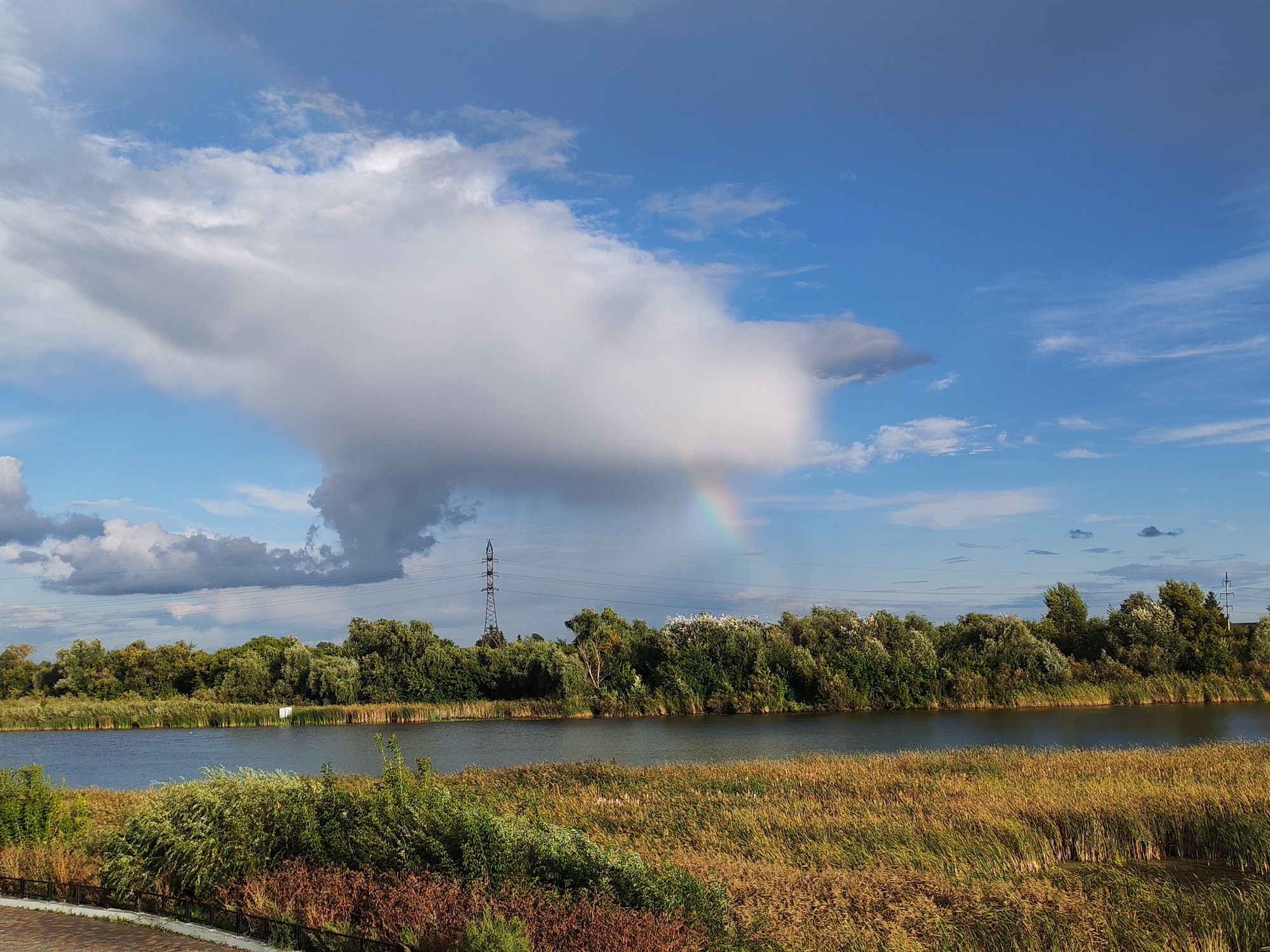The image captures a tranquil landscape scene in the city of Kurgan, Russia, likely near a body of water. A vast blue sky dominates the upper portion of the frame, adorned with scattered white and grey clouds. A notable feature is a large, cumulus cloud formation on the left, from which a faint rainbow arc emerges, suggesting recent or ongoing rainfall. Below the sky, a dense line of lush green trees forms a natural border along the far bank of a dark, calm river. The foreground features a field of tall, dry reeds and grasses, their golden-brown hues contrasting with the verdant trees and the blue water. A metal fence runs along the bottom edge of the image, separating the foreground vegetation from a paved walkway. In the distance, a tall electrical pylon stands as a solitary industrial element against the natural backdrop, with power lines extending across the scene. The overall atmosphere is serene and picturesque, highlighting the natural beauty of the Kurgan region.
