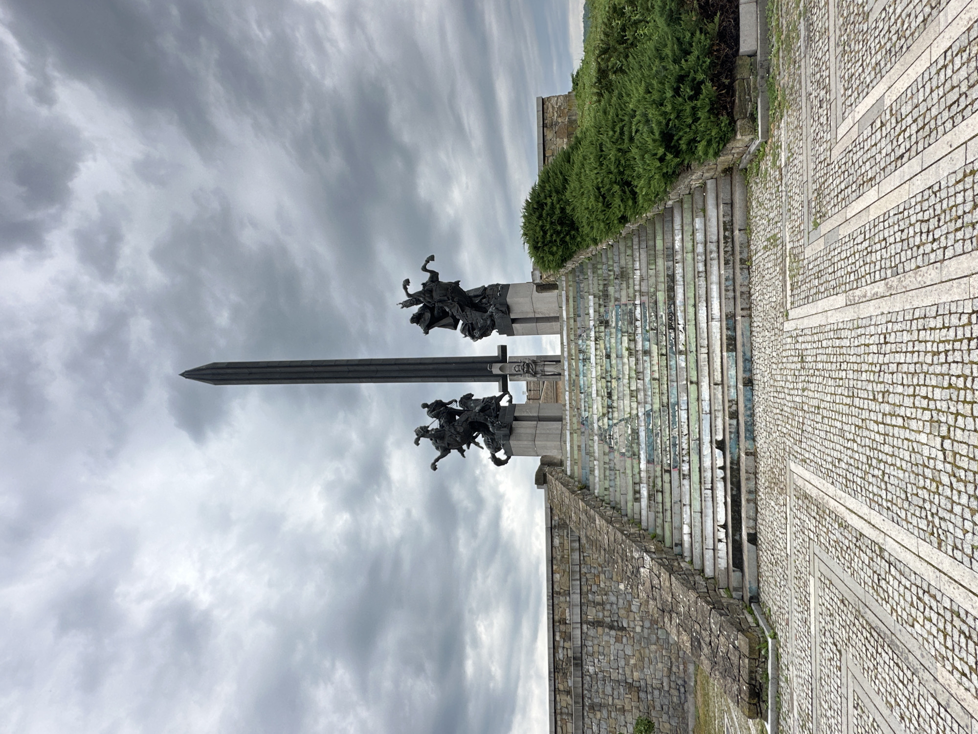The image shows the Asenevtsi Monument in Veliko Tarnovo, Bulgaria. The monument features a large sword pointing upwards, flanked by two bronze sculptures depicting figures on horseback. The sky behind the monument is cloudy and overcast. The monument is situated on a stone platform, with steps leading up to it. The surrounding area is paved with cobblestones and lined with greenery. The image is taken from a low angle, emphasizing the height of the monument.