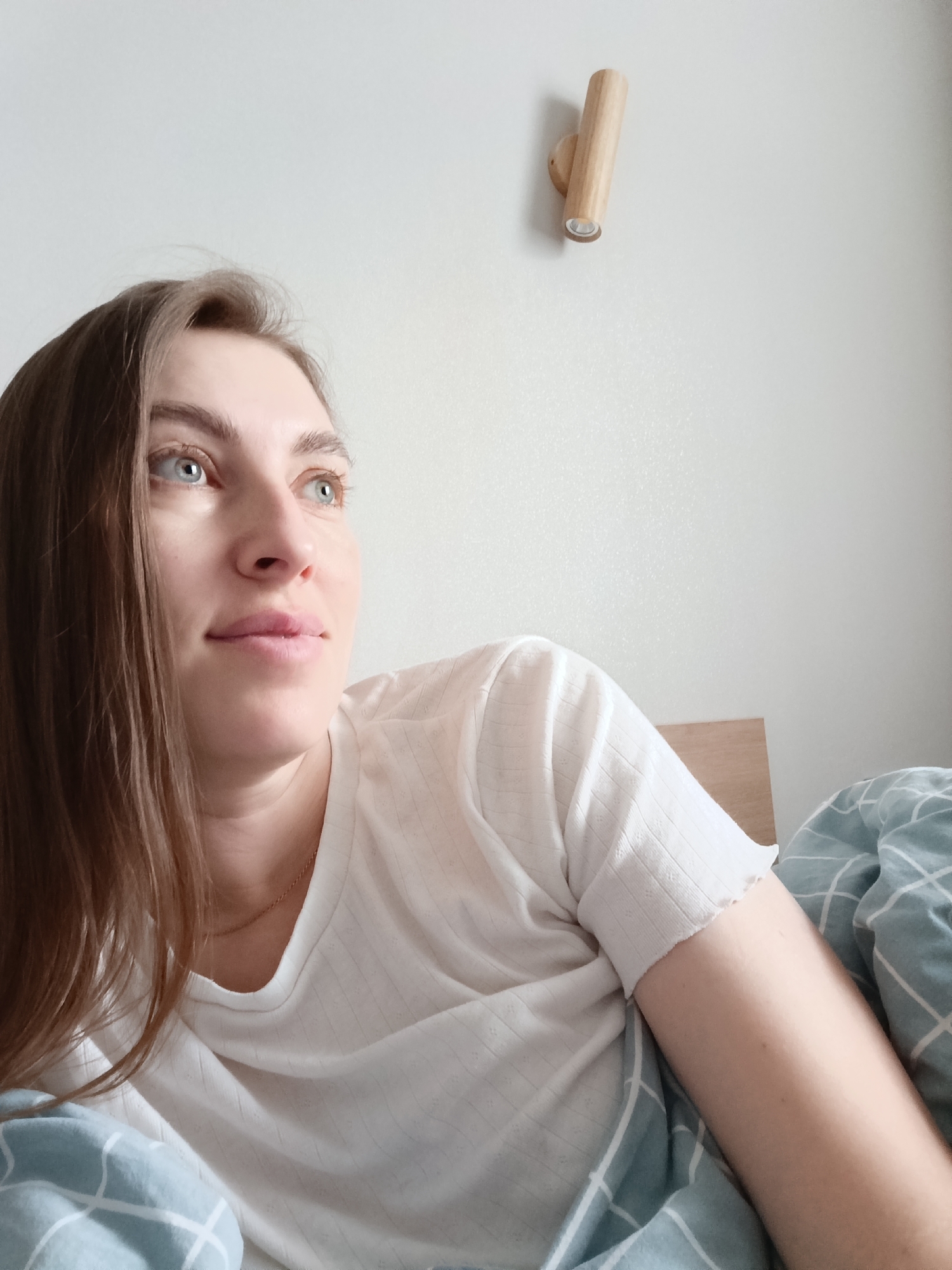 A woman with long brown hair and blue eyes is lying in bed, looking up and to the right. She is wearing a white t-shirt and a gold necklace. She is under blue and white patterned bedding. The background is a plain white wall with a wooden wall-mounted light fixture. The location is unknown but possibly in a home. The city of Chaikovsky, Russia, is not evidenced in the image.