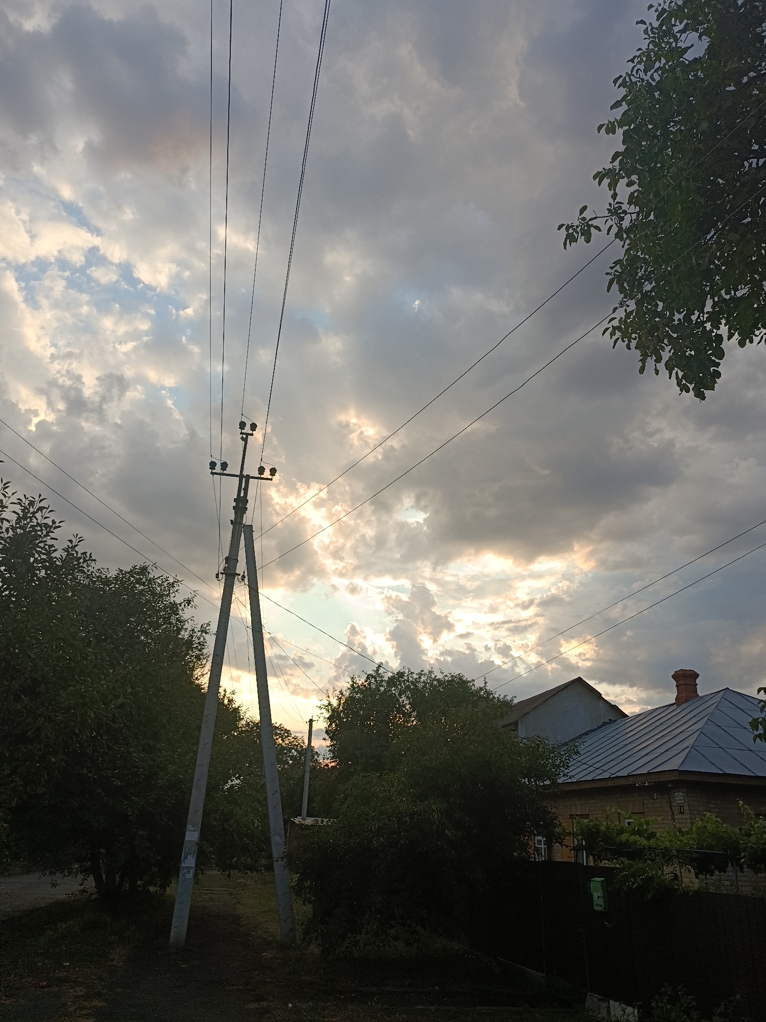 The image captures a view of a street in Smila, Ukraine, dominated by a dramatic sky filled with dark and light clouds, with rays of sunlight breaking through. In the foreground, a tall, concrete utility pole with multiple crossbars and insulators supports overhead power lines that stretch across the frame. Dense green trees flank the left side of the image, and the branches of another tree hang from the upper right corner. In the background, a residential building with a dark grey, gabled roof and a brick chimney is visible, along with parts of another structure with a lighter colored roof. A dark fence runs along the property line. The overall impression is one of a partly overcast evening in a suburban or rural setting within the city.