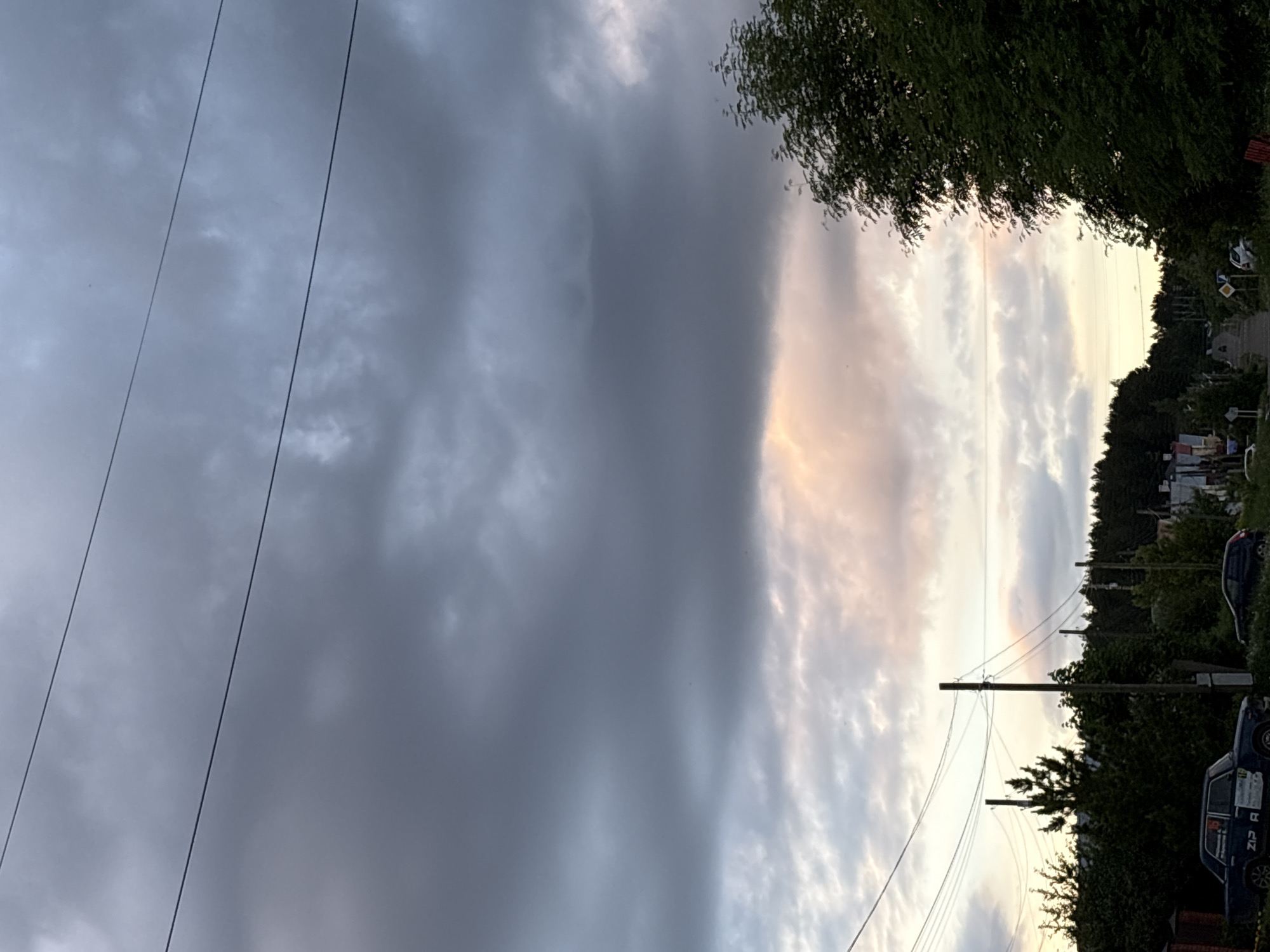 The image depicts a street scene in Прикамский, Russia. The sky is overcast with a distinct line separating dark, heavy storm clouds on the left and lighter, sunlit clouds on the right. Electrical wires run across the sky from the left side of the frame towards a power pole on the right. The foreground on the right side of the image shows trees, shrubs, and a row of buildings. Two parked vehicles are visible along the roadside. The scene is overall quiet and appears to be on the edge of a storm.
