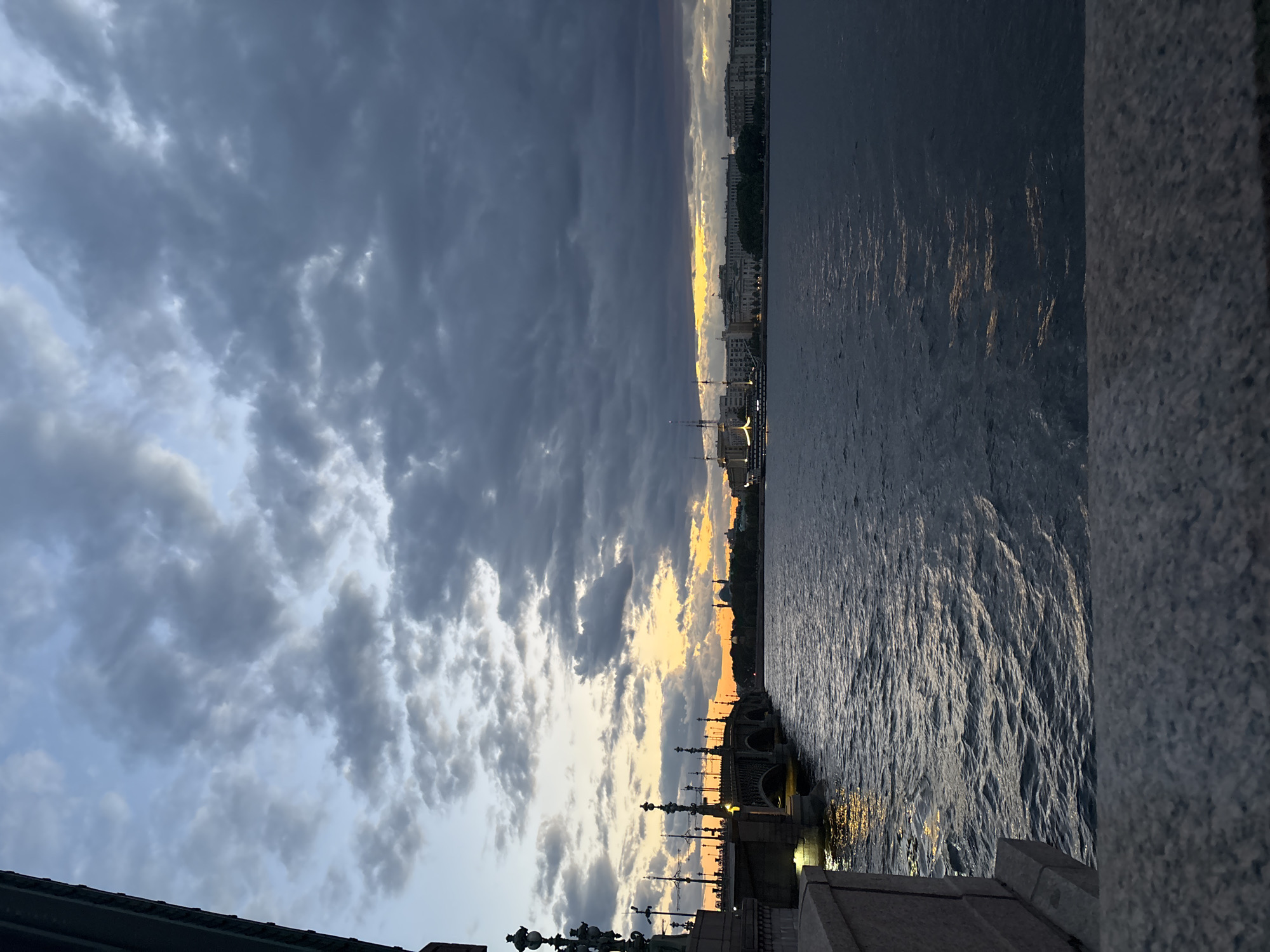The image depicts a twilight view of the Neva River in Saint Petersburg, Russia. On the left, a granite embankment and part of a bridge are visible, adorned with ornate lampposts. The bridge, an arch bridge, spans the river, with lights illuminating its structure and surrounding area. In the distance, the cityscape of Saint Petersburg unfolds, featuring buildings and a tall mast, likely from a ship. The sky is filled with dramatic, dark clouds, but a sliver of the setting sun casts a warm orange glow on the horizon, creating a striking contrast with the somber sky and deep blue water. The water's surface is rippled, reflecting the faint light. There are no people or animals visible in the image. The overall atmosphere is one of serene, dramatic beauty as day transitions into night.