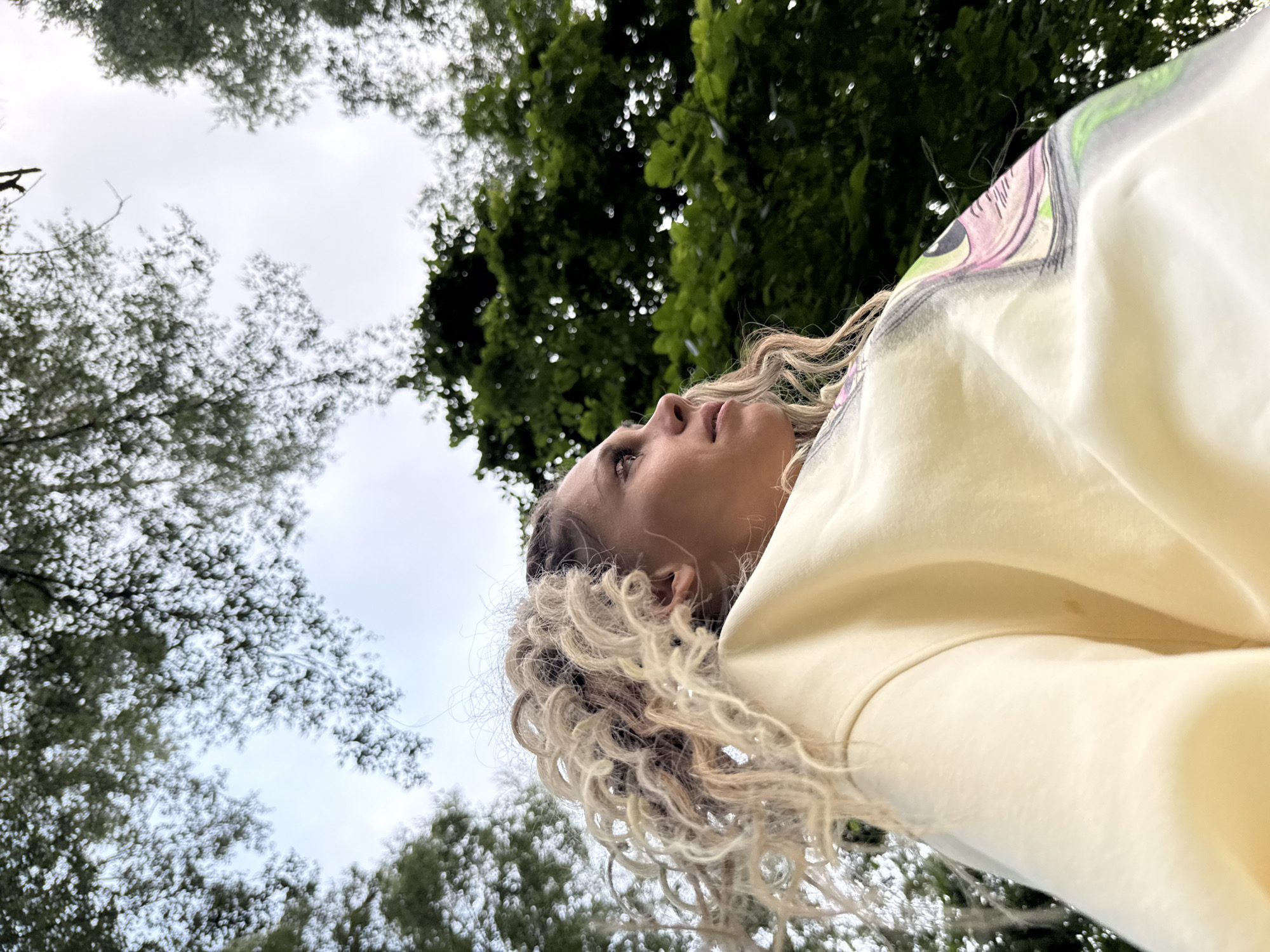 The image is a low-angle shot of a woman with blonde curly hair, wearing a light yellow sweatshirt with a graphic print, looking upwards at the trees. The background consists of green foliage and a cloudy sky. The woman's face is partially visible in profile, with her gaze directed towards the upper right of the frame. The overall scene suggests an outdoor setting, possibly a park or forest, in Rusanovo, Russia, though no specific landmarks or indications of this location are present in the image itself. The lighting is soft and diffused, consistent with an overcast day.