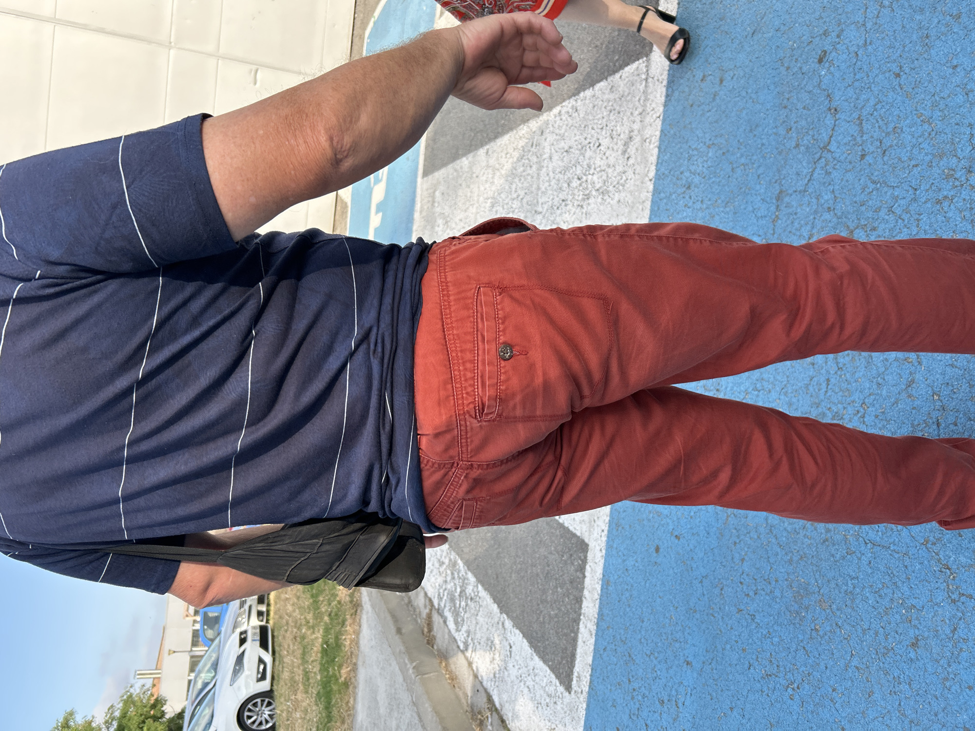 An individual wearing a navy blue horizontally striped t-shirt and red trousers stands on a street in Cerdanyola del Vallès, Spain. The ground is a textured surface with blue and white painted markings, indicating a crosswalk. In the background, several parked cars are visible, including a white sedan and a blue vehicle. The individual is partially obscured from view from the waist up, with a black bag slung over their left shoulder. A portion of another person's legs and feet, wearing black high-heeled sandals, are visible to the right of the frame. The lighting suggests it is daytime, with shadows cast on the ground.