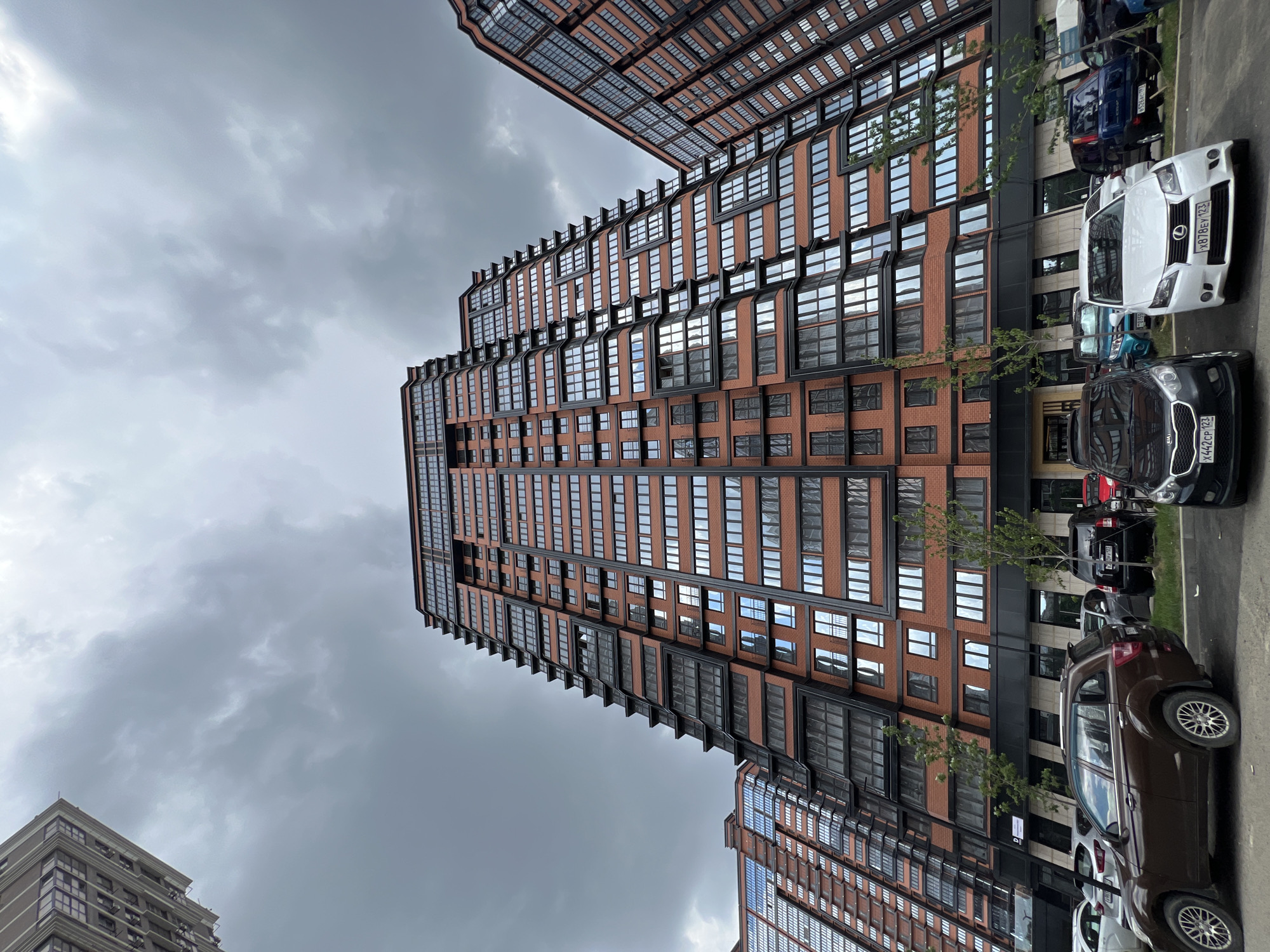 The image shows a street-level view of a modern, multi-story residential building with a brick facade and numerous windows. Several cars are parked along the street in front of the building. The sky is overcast and appears to be cloudy. The image is oriented vertically, with the building stretching upwards and the sky occupying the upper portion of the frame. The scene is set in Krasnodar, Russia. No people are visible in the image. The cars are parked, suggesting a typical day in a residential area. The lighting indicates it is daytime, possibly late afternoon, given the overcast conditions. There are no discernible emotions conveyed. Some of the car license plates are partially visible but the text is not clear enough to read.