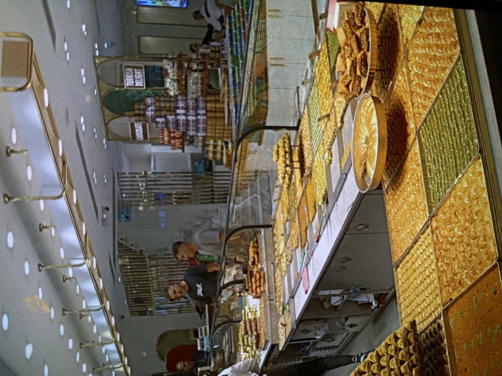 This image captures the interior of a well-lit sweets shop, likely a patisserie or confectionery, located in Bankya, Bulgaria. The primary subjects are the extensive display of golden-hued pastries, mostly baklava, arranged in large, shallow trays along a long counter, and several individuals inside the shop.
The scene depicts a bustling retail environment where customers are browsing the selection of sweets, and staff members appear to be working. In the foreground and midground, two men are visible near the display counters, one wearing a black t-shirt with "Jyld" written on it, and another in a lighter shirt. Another person in a white uniform can be seen further back, possibly a staff member.
Notable details include the sheer abundance and variety of the pastries, which are neatly arranged in patterns within the trays. Some trays feature diamond-shaped pieces, others intricate rolls, and some appear to be different types of cakes or desserts, including a green-colored variety. Two large, round, golden serving dishes are also present on the main counter. Beyond the main pastry counter, shelves stocked with packaged goods are visible. Signs above these shelves clearly display "TURKISH DELIGHT" and "CHOCOLATE", indicating other items sold. The shop features bright overhead lighting with numerous recessed lights in the ceiling, creating an inviting atmosphere. The overall impression is one of a specialty store offering a wide array of sweet treats. The time of day appears to be daytime, given the general illumination, though it is an indoor setting. No strong emotional cues are evident, but the scene suggests a typical retail interaction.