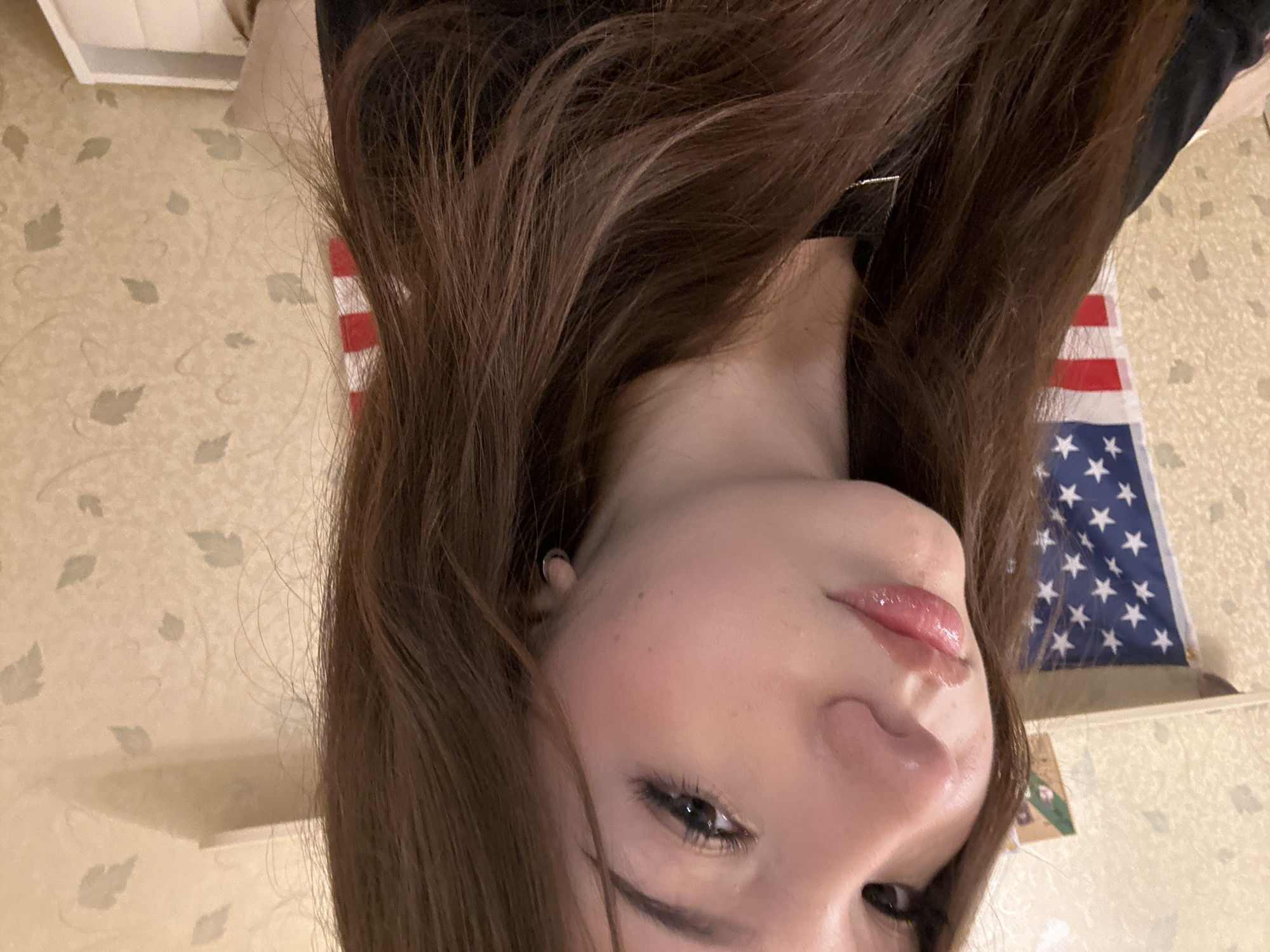 The image displays a close-up, portrait-style view of a young person, likely female, with fair skin and long, wavy dark brown hair. The person's face is prominent, featuring defined eyebrows, visible eyelashes, and glossy, pink-toned lips slightly parted. A subtle blush is visible on the cheeks. A small, circular earring is present in the visible left ear. The individual wears a black garment and a thin silver necklace.
The background on the left side features a partially visible American flag, displaying white stars on a blue field and red and white stripes. Above and behind the flag is a light-colored, possibly textured wall, with a narrow shelf running horizontally. A small, dark-colored object rests on this shelf. The right side of the background reveals a light-colored wall adorned with a recurring pattern of faded, grey-green leaf motifs. Another shelf is visible in the upper right. In the lower right corner, there is a light-colored soft object, possibly a cushion, next to a white rectangular container or tray. This scene is stated to be located in the city of Naberezhnye Chelny, Russia.