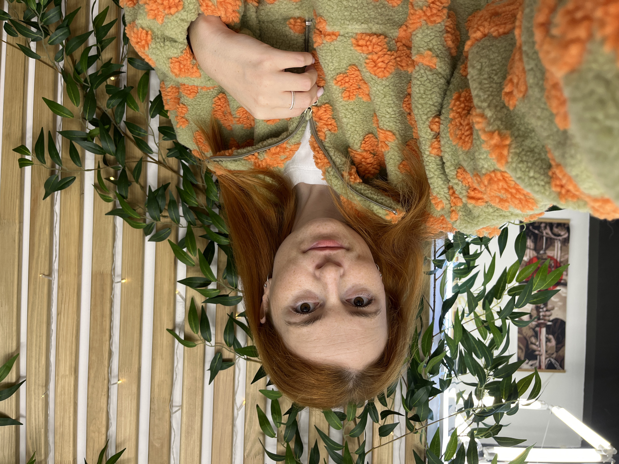 A red-haired woman with fair skin and freckles is captured in a medium close-up selfie. She is wearing a light green fuzzy jacket with an orange floral pattern, partially zipped. A white t-shirt is visible underneath the jacket. She has on a thin silver ring on her left hand, which is resting near the zipper. She is wearing small hoop earrings. The background consists of artificial green foliage intertwined with thin strings of fairy lights, set against a wall of vertical wooden planks. To the left of the woman, a portion of a dark wall is visible, with a framed artwork hanging on it. The scene appears to be indoors, possibly in a home or a business, and is not specifically identifiable as being in the city of Chaikovsky, Russia.