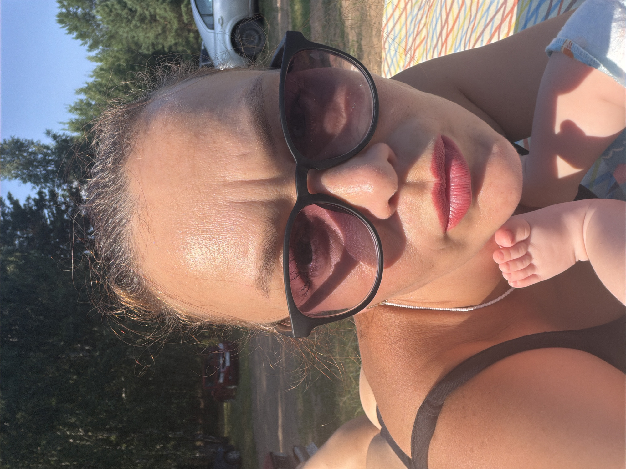 This media file is a close-up, vertical, selfie-style photograph featuring two individuals, an adult and an infant, outdoors on a sunny day. The adult is wearing sunglasses and has bright red lipstick on. The infant's foot is visible in the upper right portion of the frame, resting on the adult's chest. The adult is wearing a black bikini top and a white beaded necklace. In the background, blurred trees and a vehicle can be seen, suggesting a park or rural setting. The lighting indicates it is daytime, with the sun casting shadows. The overall mood appears relaxed and affectionate. There is no visible text in the image.