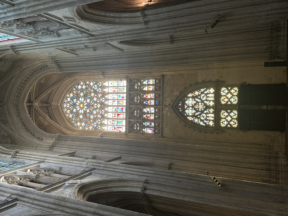 The image shows the interior of the Saint-Gatien Cathedral, located in Tours, France, at the coordinates 47.395 latitude and 0.694 longitude. The cathedral features a large, arched stained glass window, comprised of three sections, and is set within a high, vaulted interior. Sunlight streams through the window, illuminating the interior with colorful light. There are no people in the image. The window is comprised of several panels of stained glass, depicting various scenes and motifs. The floor is covered in stone tiles. The walls are made of stone and are adorned with decorative carvings. The overall appearance of the cathedral is one of grandeur and solemnity.