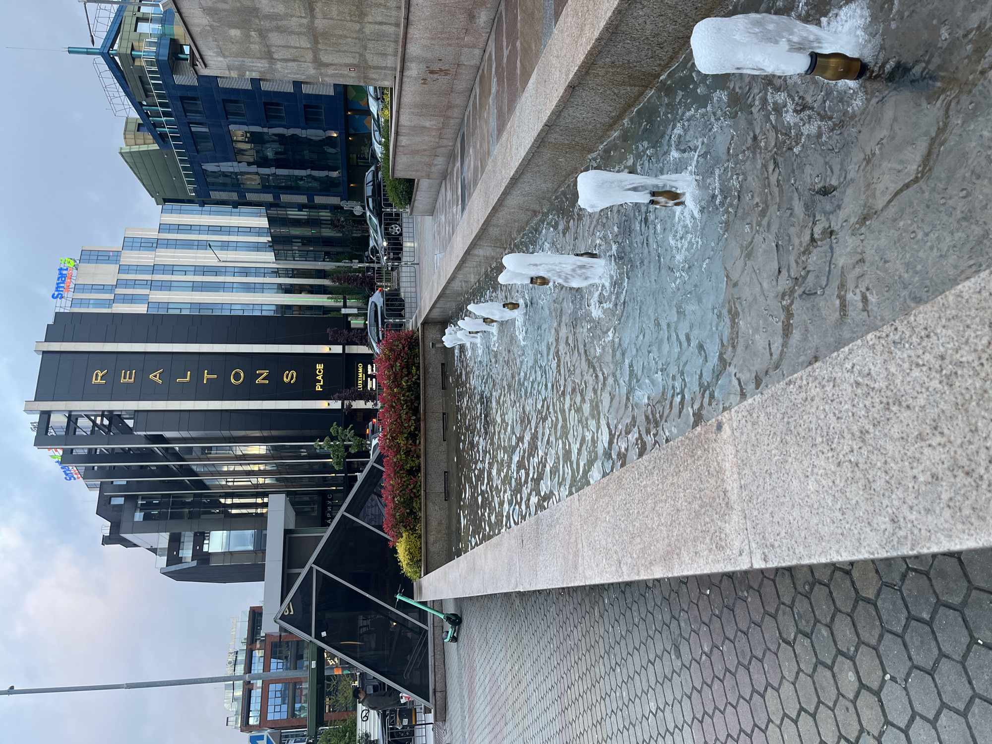 This image depicts a modern urban scene in Sofia, Bulgaria, characterized by contemporary architecture and a public water feature. In the foreground, a long, rectangular fountain with a light-colored, speckled stone basin is active, featuring multiple brass nozzles spraying white, aerated jets of water.
Adjacent to the fountain, a paved walkway is visible, made of dark grey hexagonal tiles. A bright green electric scooter stands parked on this paving, suggesting urban transportation. Next to the fountain, a planter filled with red and green ornamental bushes adds a touch of color to the concrete and glass environment.
The background is dominated by several multi-story buildings. A prominent structure on the left features a dark facade with large, gold letters spelling "REALTONS PLACE" vertically. Above this, the "Smart" logo (likely "Smart Office" or "Smart Living") is visible on a building with a lighter, blue and white glass facade. Further behind and to the right, a dark blue building with numerous reflective windows and a distinctive angular rooftop structure adds to the modern skyline.
Other elements include several parked cars, partially obscured by a metal barrier, and a street light pole. A glass-paneled structure with a partially visible Cyrillic word "АРИУС" (ARIUS) is also present. The sky is overcast, indicating a cloudy day, and the lighting suggests daylight, possibly late afternoon. The scene portrays a functional and aesthetically pleasing commercial or business district. Small text "EUXIMMO" is also visible near "PLACE".