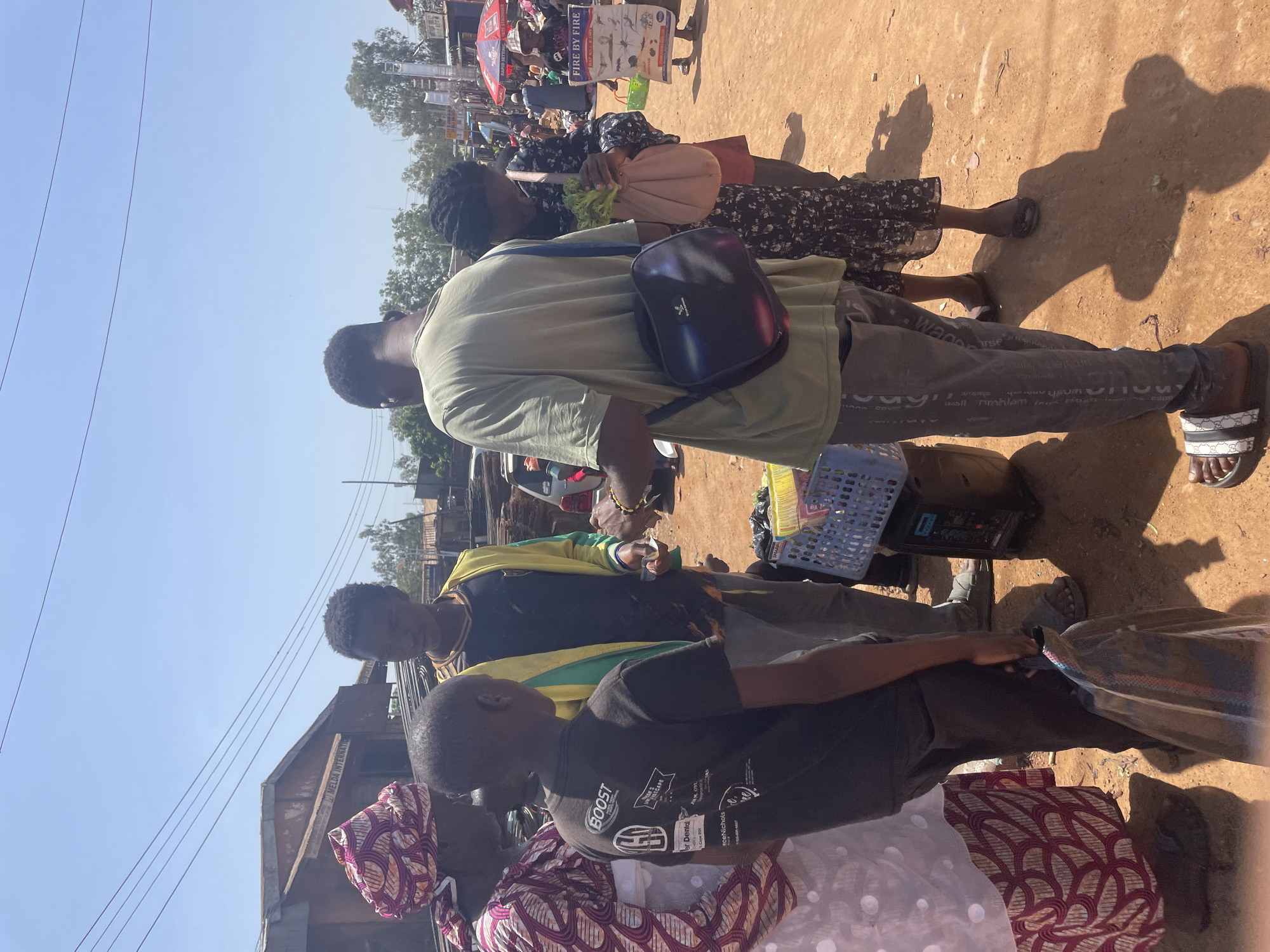 This full-shot image, taken in Gura Top, Nigeria, captures a bustling outdoor street market or pathway under bright daylight. The ground is reddish-brown dirt, and the sky is a clear, light blue, with power lines visible overhead.
Numerous individuals are present, engaged in various activities typical of a market setting. In the mid-ground, a man in a light olive-green t-shirt and dark pants with white script text (including "WADE", "problem", "wall") stands, wearing a dark shoulder bag with a distinct logo. He wears white-patterned slides on his feet. Behind him, a woman in a dark floral dress carries a light-colored fabric bag, likely containing green produce. Strong, distinct shadows are cast on the ground, indicating bright, direct sunlight, suggesting a time around midday.
Towards the left foreground, another man in a black shirt with a yellow and green patterned garment draped over his shoulders looks towards the right. He appears to be interacting with a younger person wearing a black t-shirt that prominently displays the text "BOOST," "Dental," "ceNichols," and "SCAPE." Partially visible beside them is an older woman in a vibrant, patterned headscarf and dress.
Objects in the scene include a blue plastic basket and a black electronic device near the feet of the man in the green shirt. In the background, market stalls are visible, along with a sign that clearly reads "FIRE BY FIRE" and "RAT KILL," suggesting the sale of pest control products. Another building in the background displays the text "MELDA INTERNATION." The scene conveys a lively commercial atmosphere with people shopping and interacting amidst goods and vendors.