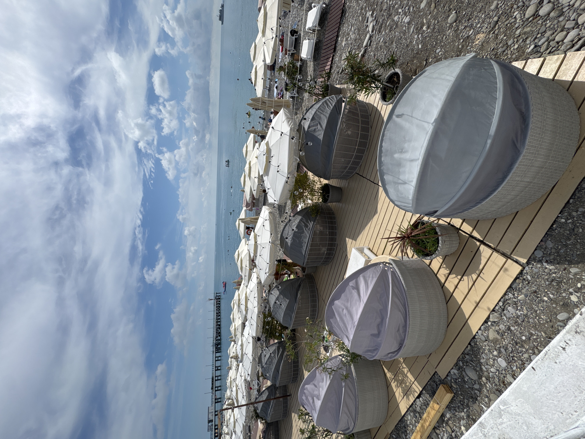 This image depicts a view of a beach resort in Sochi, Russia, on a cloudy day. In the foreground, several circular lounge chairs with grey covers are arranged on a wooden deck. Beyond the chairs, a beach with white umbrellas and sun loungers is visible. The sea is calm, and in the distance, a pier with structures and a ship can be seen. The sky is filled with scattered clouds. There are no people clearly visible in the foreground, but there are individuals in the water and on the beach in the background.