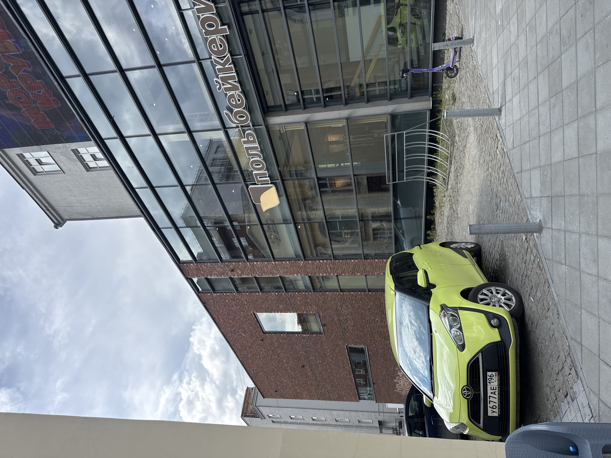 The image captures a street scene in Yekaterinburg, Russia, featuring a modern building with a brick facade and extensive glass windows. A bright lime-green Toyota Sienta minivan is parked on a cobblestone street in front of the building. Above the minivan, the building's facade displays the text "ПОЛЬ БЕЙКЕРИ" in white, stylized lettering, accompanied by a brown logo resembling a loaf of bread or a pastry. Another identical lime-green car is visible reflected in the glass facade. To the right of the minivan, a purple electric scooter stands near metal bollards. In the background, an older, light gray building with visible windows is seen. The sky is overcast with clouds.