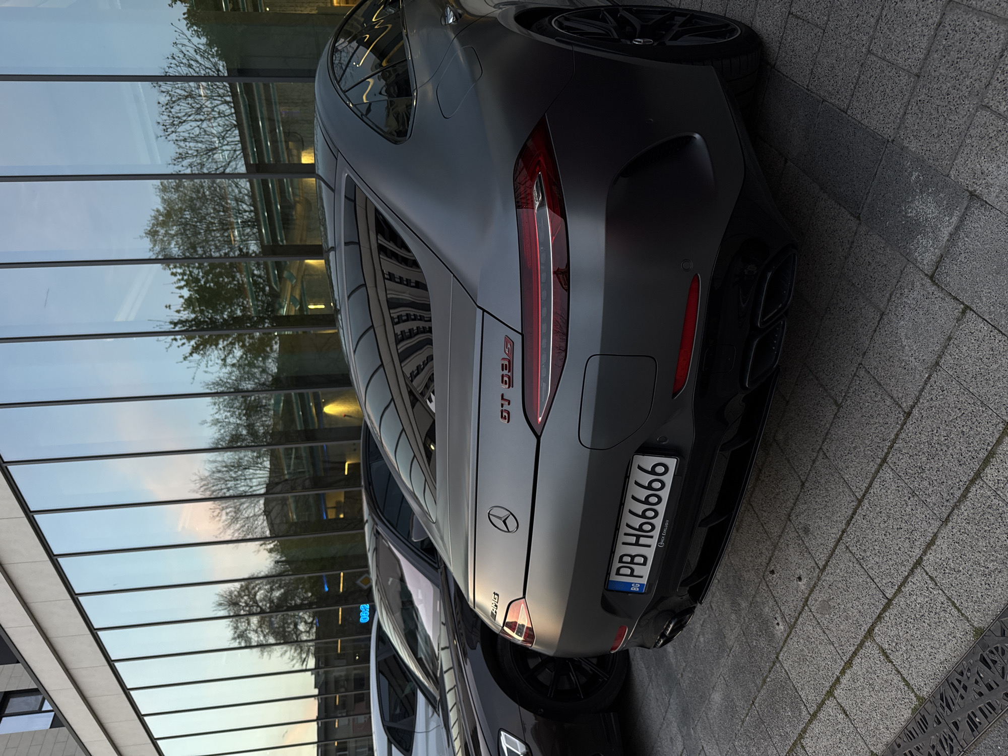 The image shows the rear of a matte gray Mercedes-AMG GT 63 S 4-Door Coupé, bearing a Bulgarian license plate "PB H66666". The car is parked on a gray stone-paved surface, with a modern building in the background featuring a reflective glass façade that mirrors the surroundings, including trees and the sky. The license plate frame has the inscription "CHIP EXHAUST". The car appears to be in Plovdiv, Bulgaria. There are no people visible in the frame, and the focus is solely on the vehicle and its immediate surroundings. The photograph's perspective is slightly tilted.