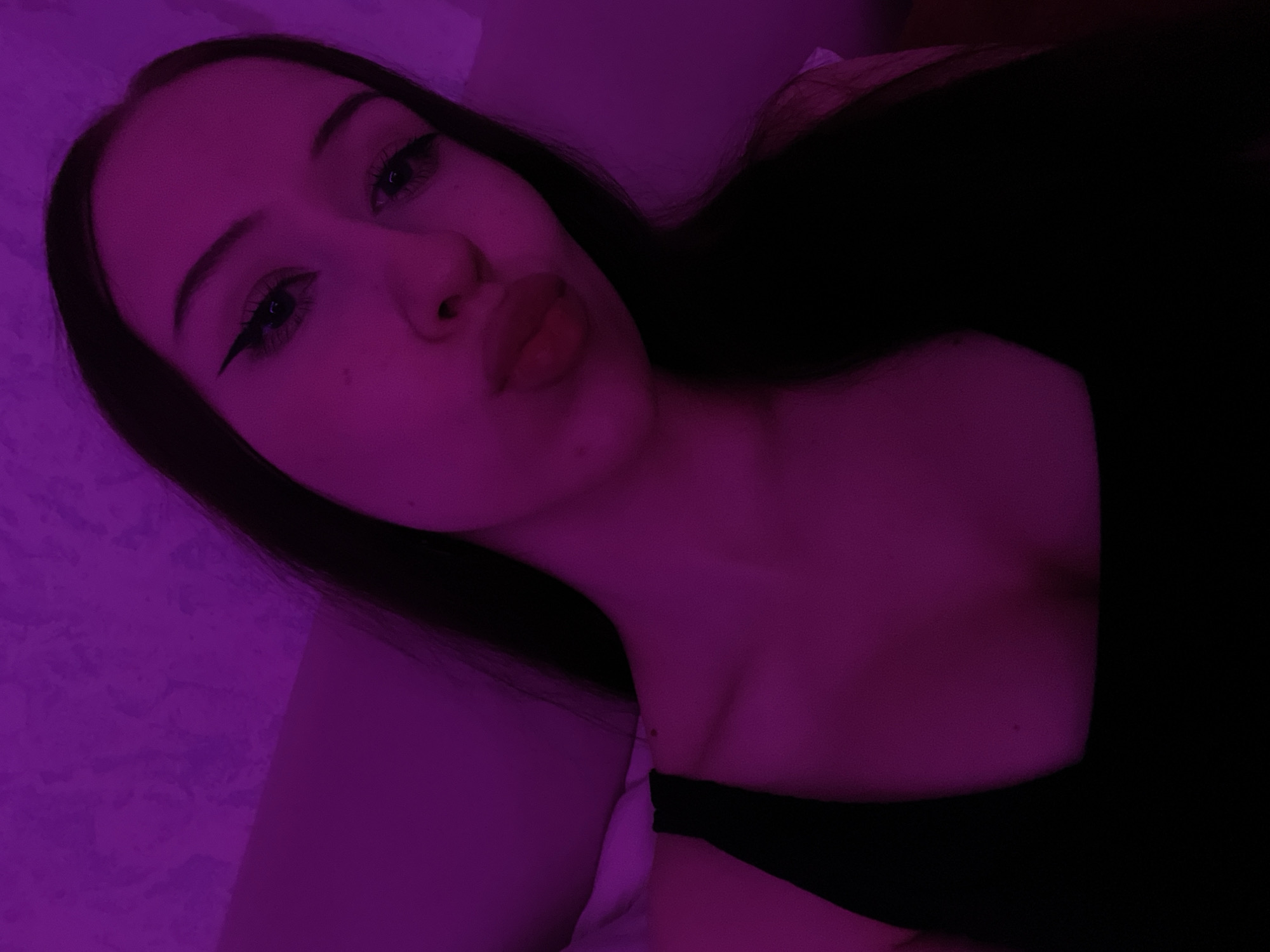The image shows a close-up, high-angle selfie of a young woman with long, dark hair. She is wearing a black top and her makeup includes winged eyeliner and defined eyebrows. Her lips are pursed. The lighting is a vibrant pinkish-purple, casting a dramatic glow on her skin and the textured background, which appears to be a wall or bedding. The scene is dimly lit, with the light source likely being artificial and positioned to create a moody ambiance. No specific location in Votkinsk, Russia, can be identified from the visual information provided.